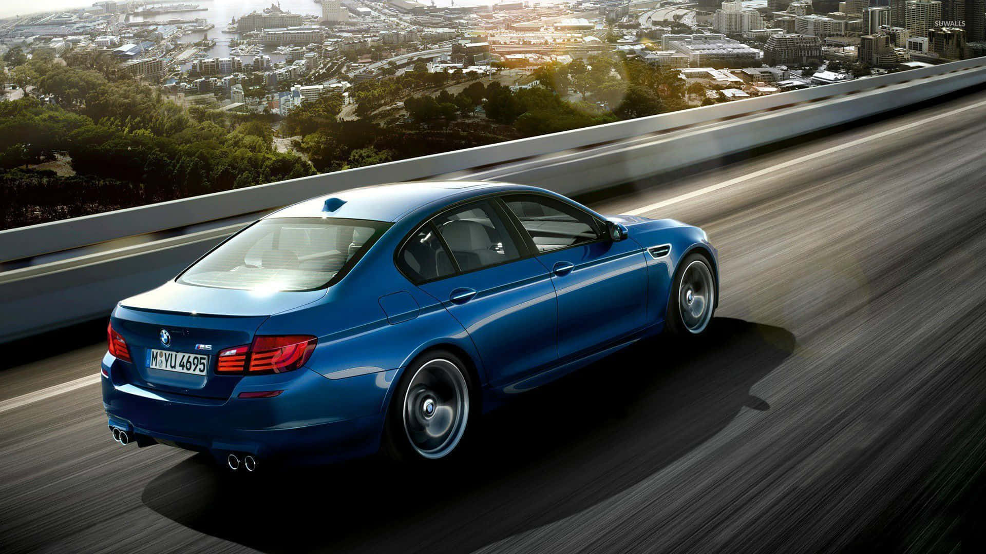 BMW 動圖 桌布 壁紙 — 一輛時尚的藍色BMW M5在高速公路上奔馳，下方是金色陽光下廣闊的城市景觀和綠地。