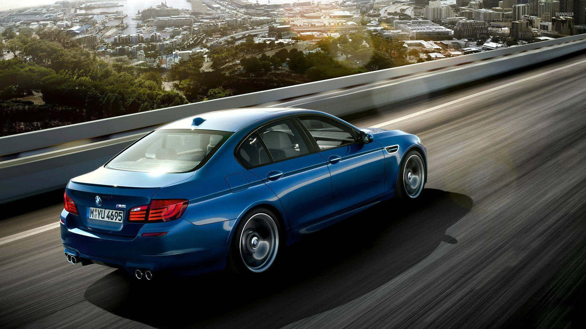 Blue Car 桌布 壁紙 — 一輛時尚的藍色BMW M5在高速公路上奔馳，下方是金色陽光下廣闊的城市景觀和綠地。