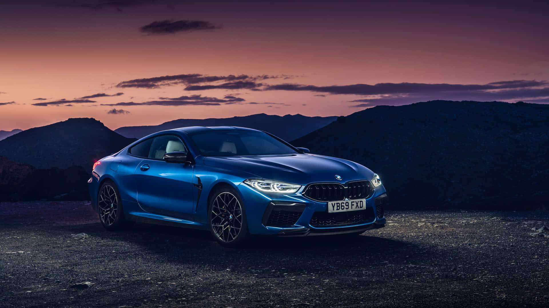 1920x1080 Bmw 桌布 壁紙 — 一輛時尚的藍色BMW M8 Coupe在暮色天空下閃耀，崎嶇山脈上方有紫色和橙色的雲彩。