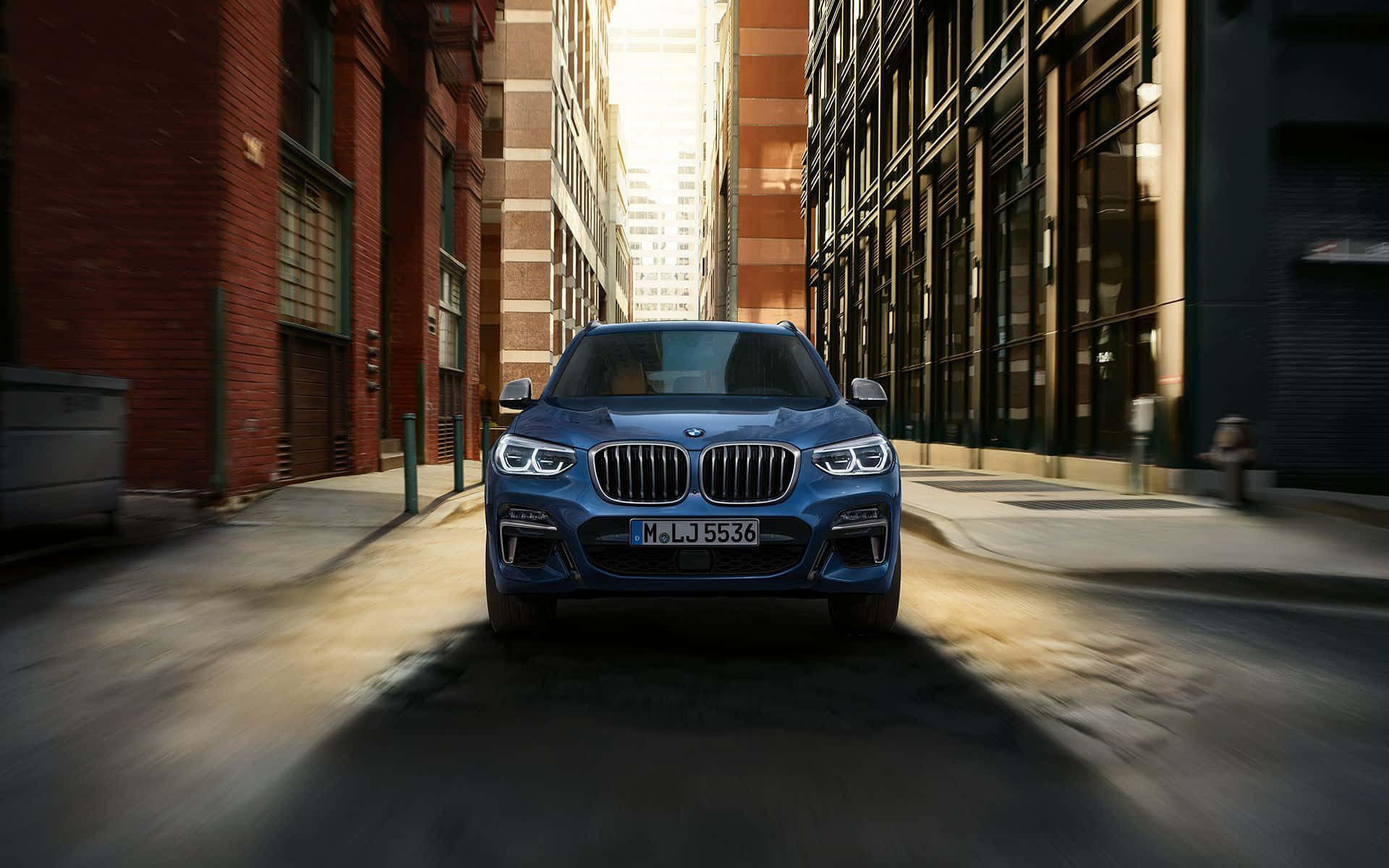 Bmw X3 桌布 壁紙 — 一輛時尚的藍色BMW SUV行駛在一條狹窄的城市小巷中，兩旁是磚砌和玻璃建築，金色的陽光穿透而過。