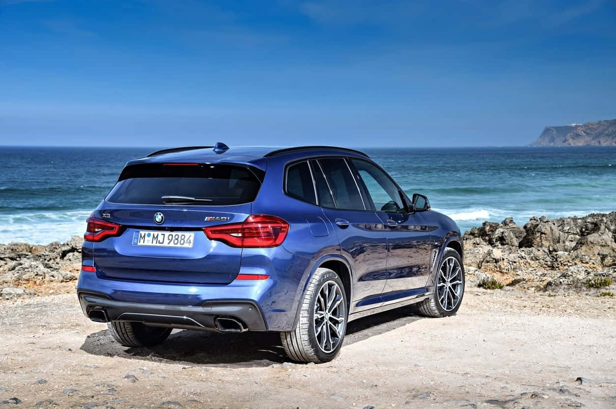 Bmw X3 桌布 壁紙 — 一輛時尚的藍色BMW X3 M40i SUV停泊在岩石海岸懸崖上，俯瞰海洋，天空湛藍清澈。
