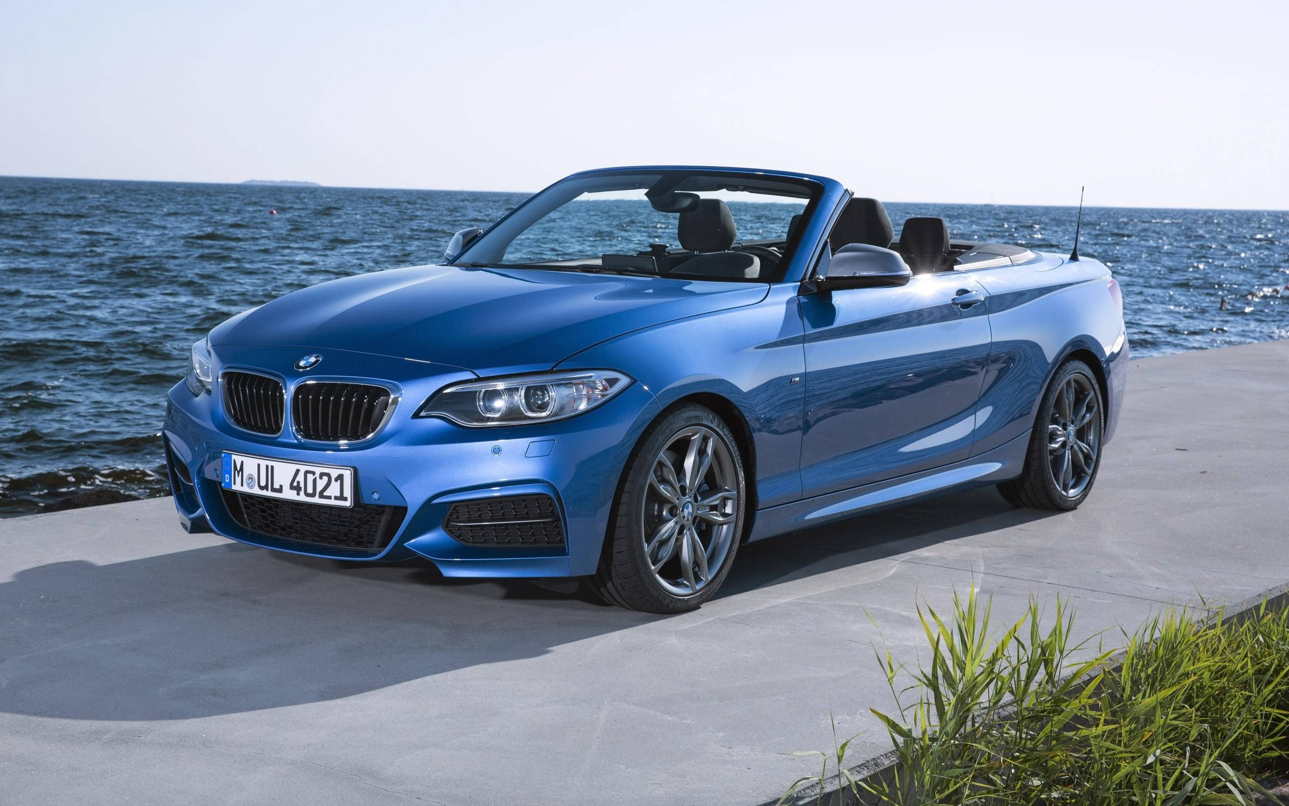 Bmw M4 桌布 壁紙 — 一輛時尚的藍色BMW敞篷車停在混凝土碼頭上，背後在明亮的日光下海浪拍打。