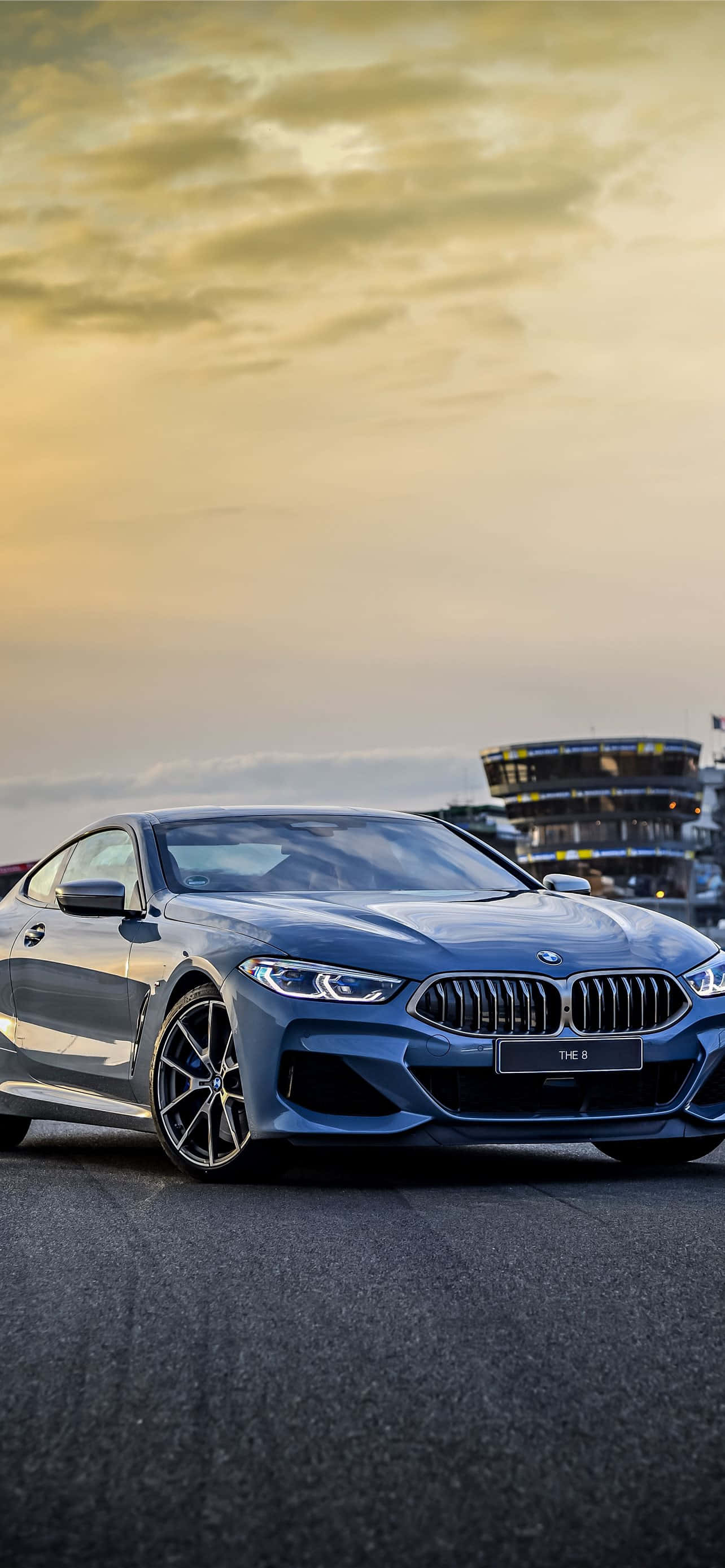 Bmw 328 桌布 壁紙 — 一輛時尚的藍色BMW跑車停在瀝青路上，背景是金色夕陽天空和賽車場看台。