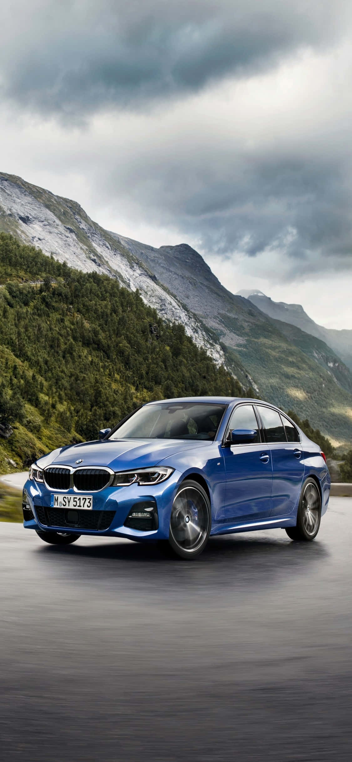 iPhone X Bmw 桌布 壁紙 — 一輛時尚的藍色BMW轎車在戲劇性的多雲天空下行駛在山路上。
