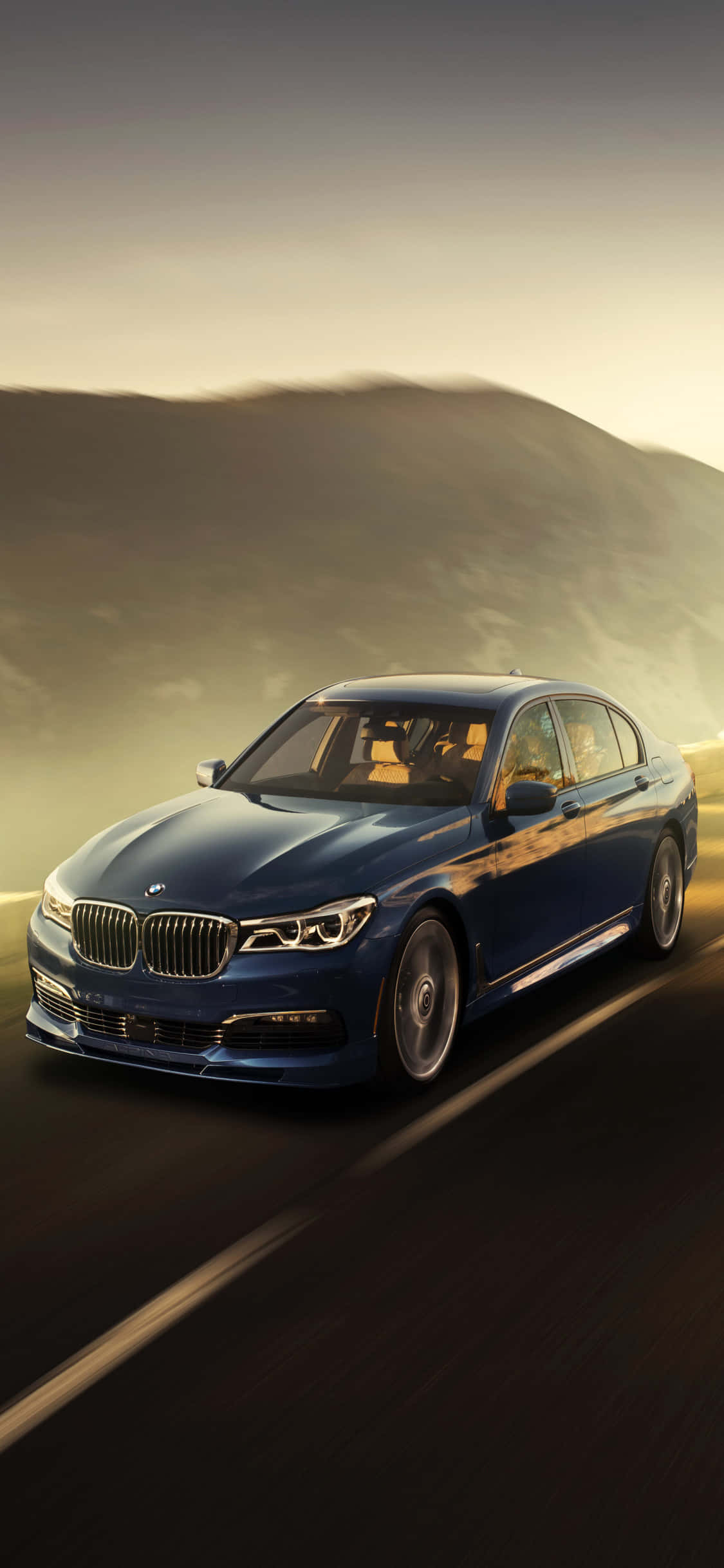 iPhone X Bmw 桌布 壁紙 — 一輛時尚的藍色BMW轎車在黃金時刻沿著蜿蜒的道路行駛，背景是朦朧的山脈。