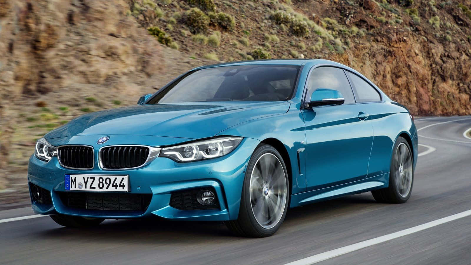 Bmw 4 Series 桌布 壁紙 — 一輛時尚的藍色BMW雙門跑車沿著蜿蜒的山路疾馳，背景是模糊的岩石地形。