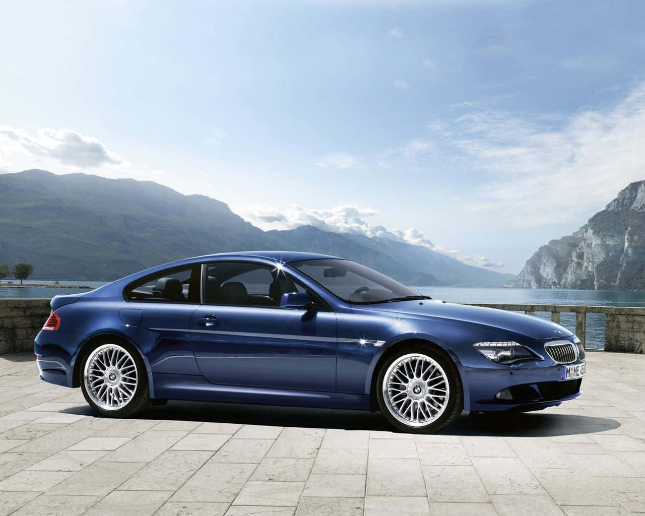 Bmw 6 Series 桌布 壁紙 — 一輛時尚的藍色BMW雙門轎跑車在石頭露台上閃閃發光，俯瞰著寧靜的湖泊，背景是山巒，天空明亮。