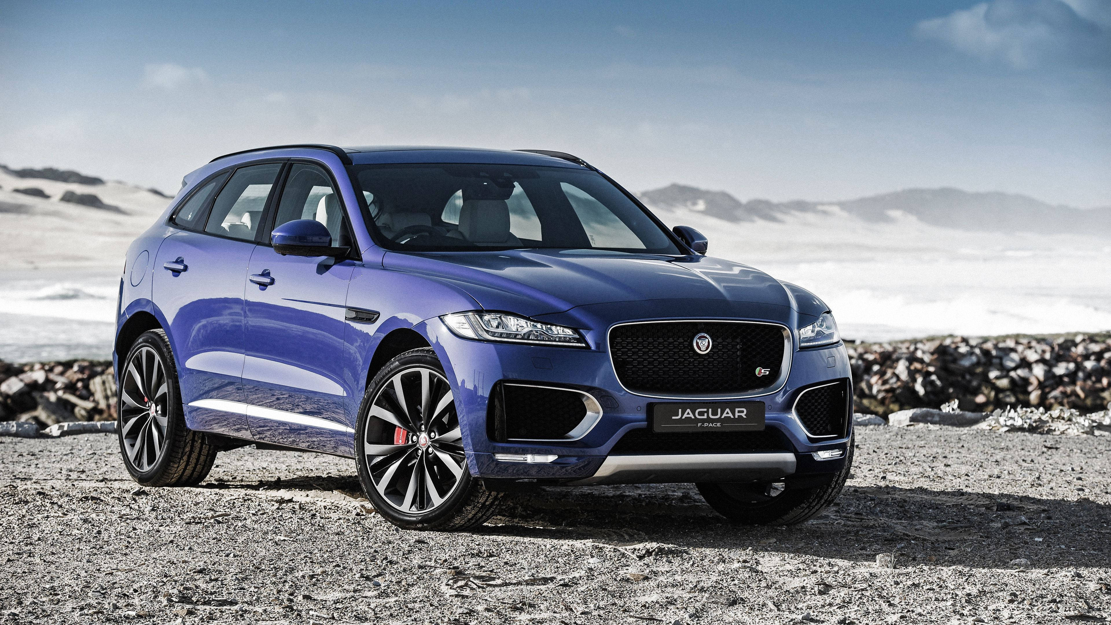 美洲豹汽車 桌布 壁紙 — 一輛時尚的藍色Jaguar F-Pace SUV停在岩石海灘上，背景是白雪覆蓋的山脈和蒼白的天空。