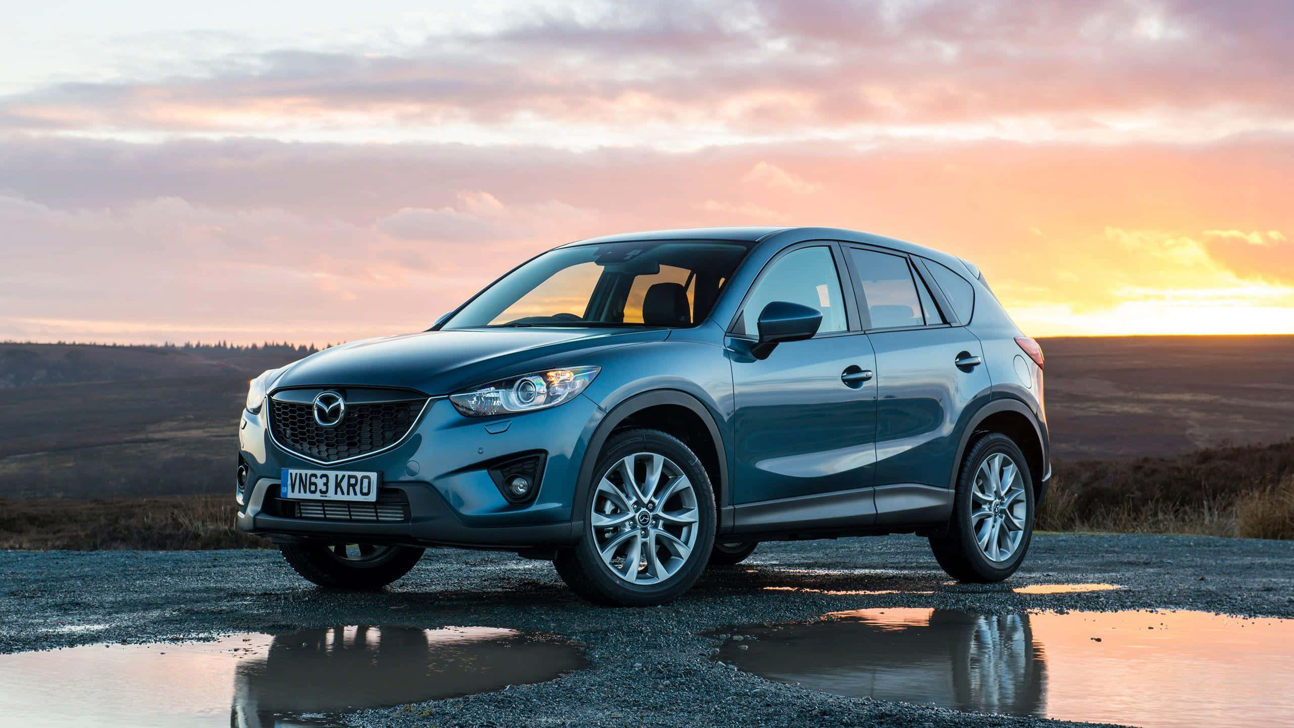 Mazda Cx-5 桌布 壁紙 — 一輛時尚的藍色Mazda CX-5 SUV停泊在日落時分的濕碎石路上，水坑倒映著天空。