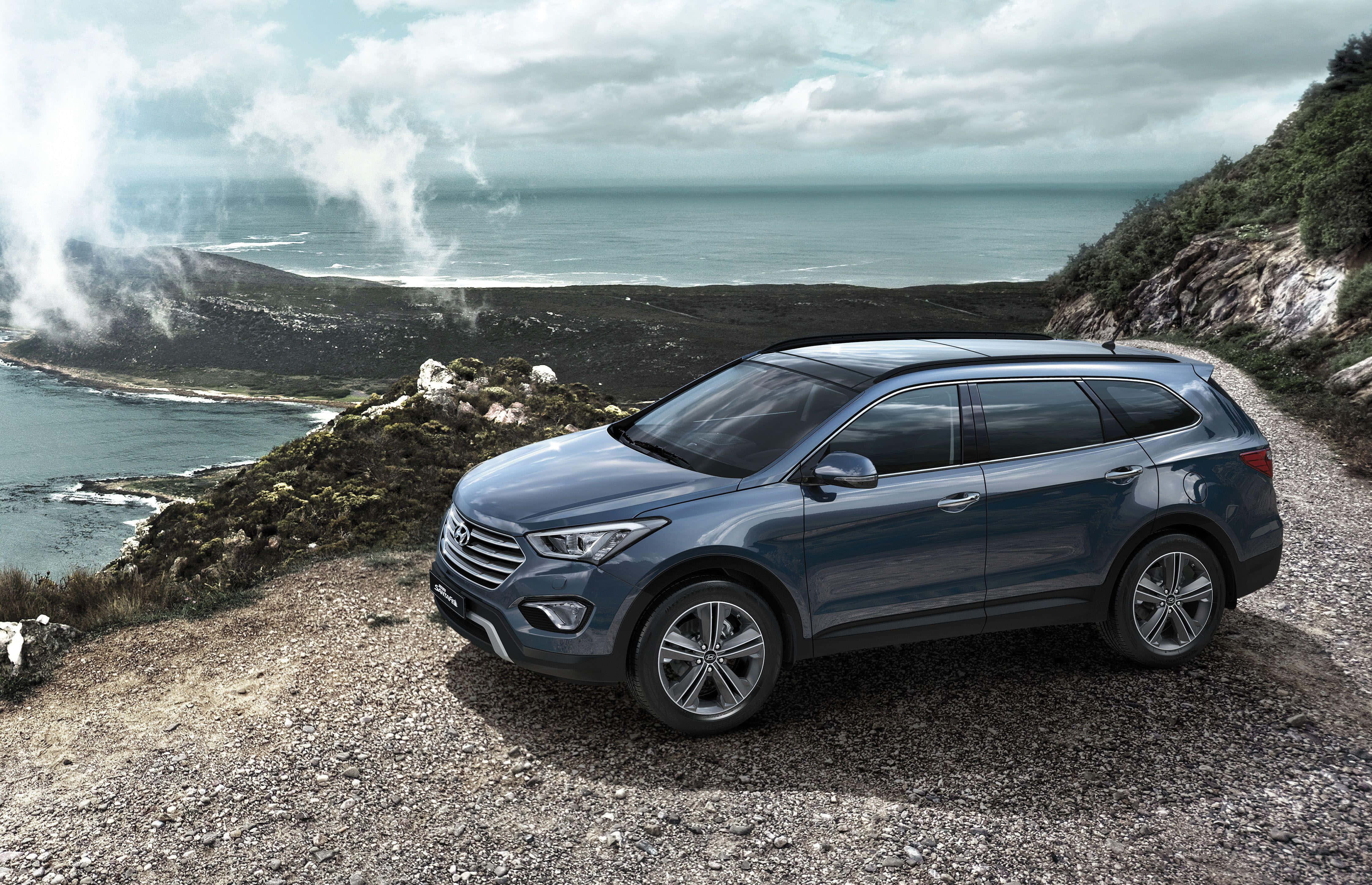 Hyundai Santa Fe 桌布 壁紙 — 一輛時尚的藍色SUV停泊在崎嶇的海岸懸崖上，俯瞰著霧氣瀰漫的海洋，上方有戲劇性的雲層。