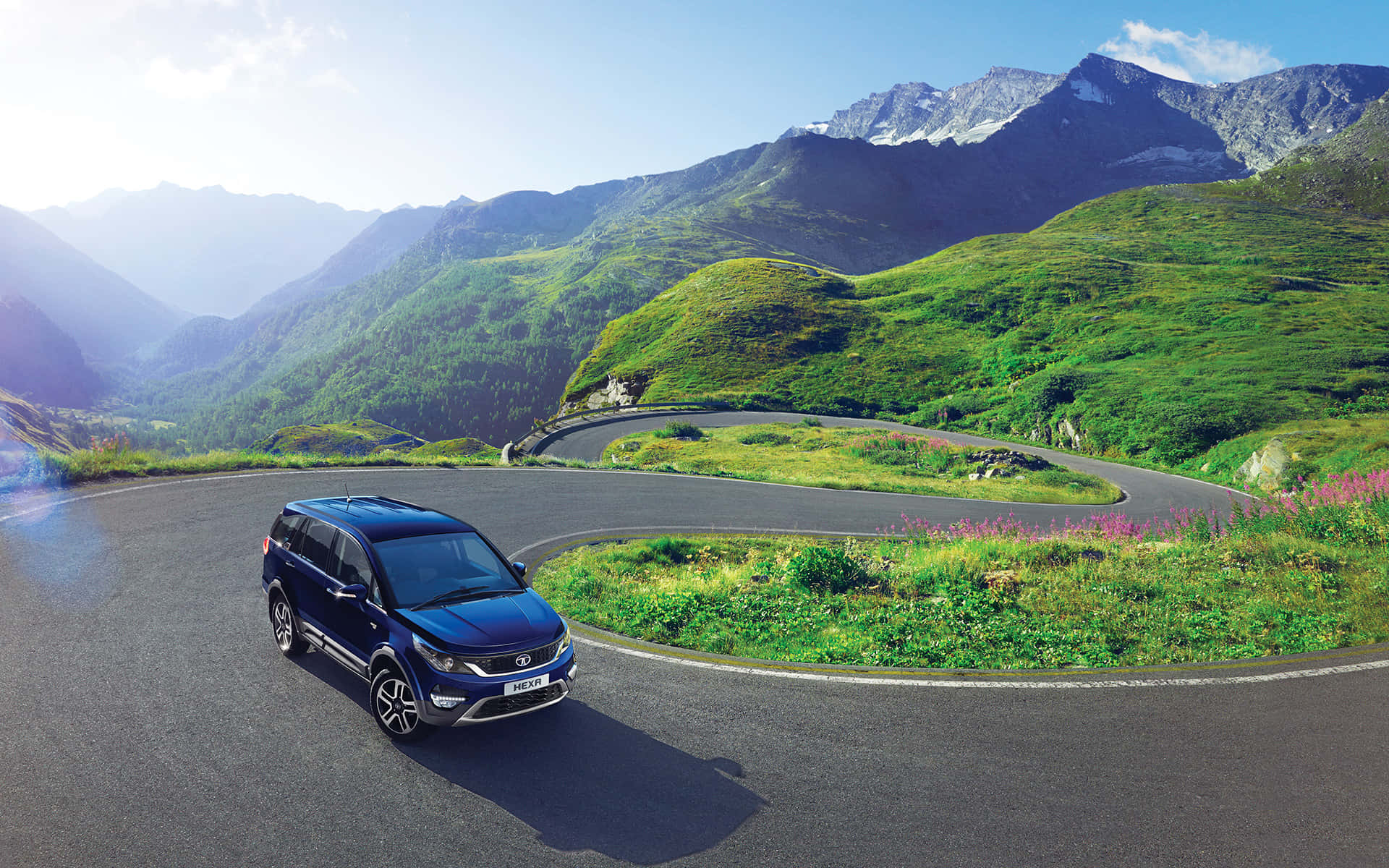 Tata Motors 桌布 壁紙 — 一輛時尚的藍色SUV在蜿蜒的山路上行駛，周圍是茂密的綠色山丘和白雪覆蓋的山峰，天空湛藍。