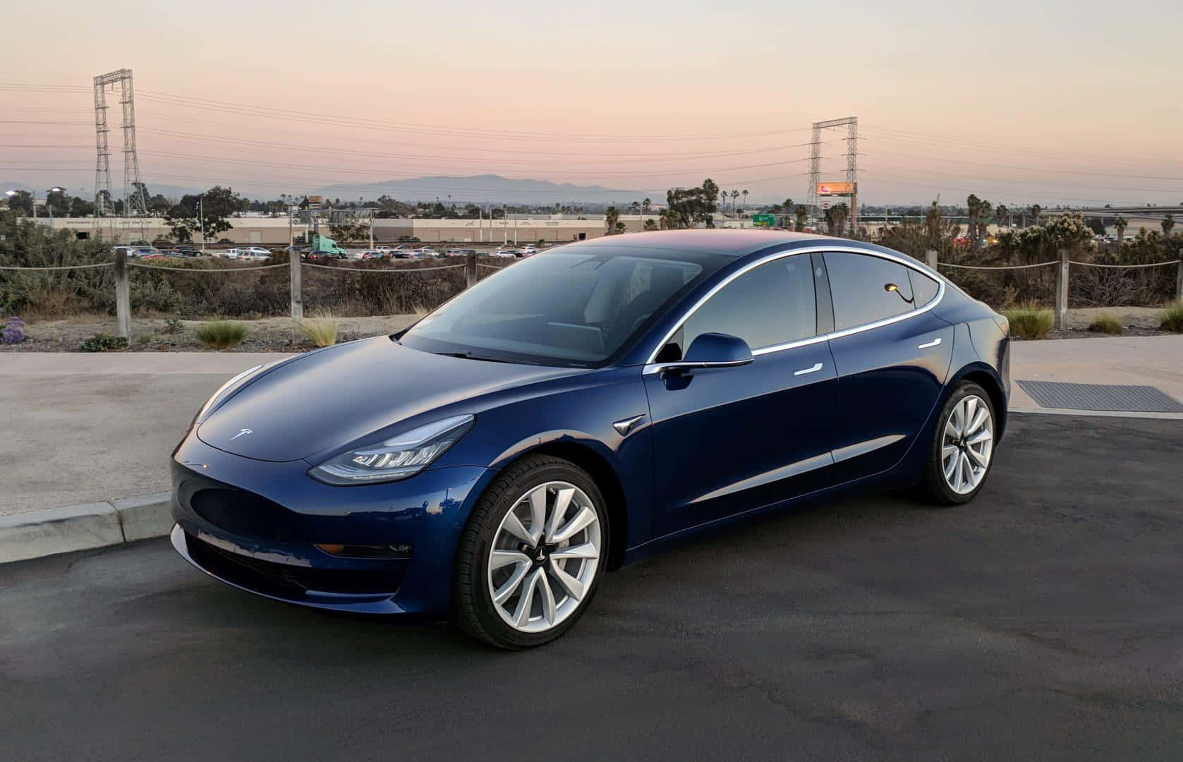 Tesla Model 3 桌布 壁紙 — 一輛時尚的藍色Tesla Model 3在黃昏時停泊在瀝青路上，背景有電線和遠處的山丘，天空呈現柔和的粉紅色和橙色。