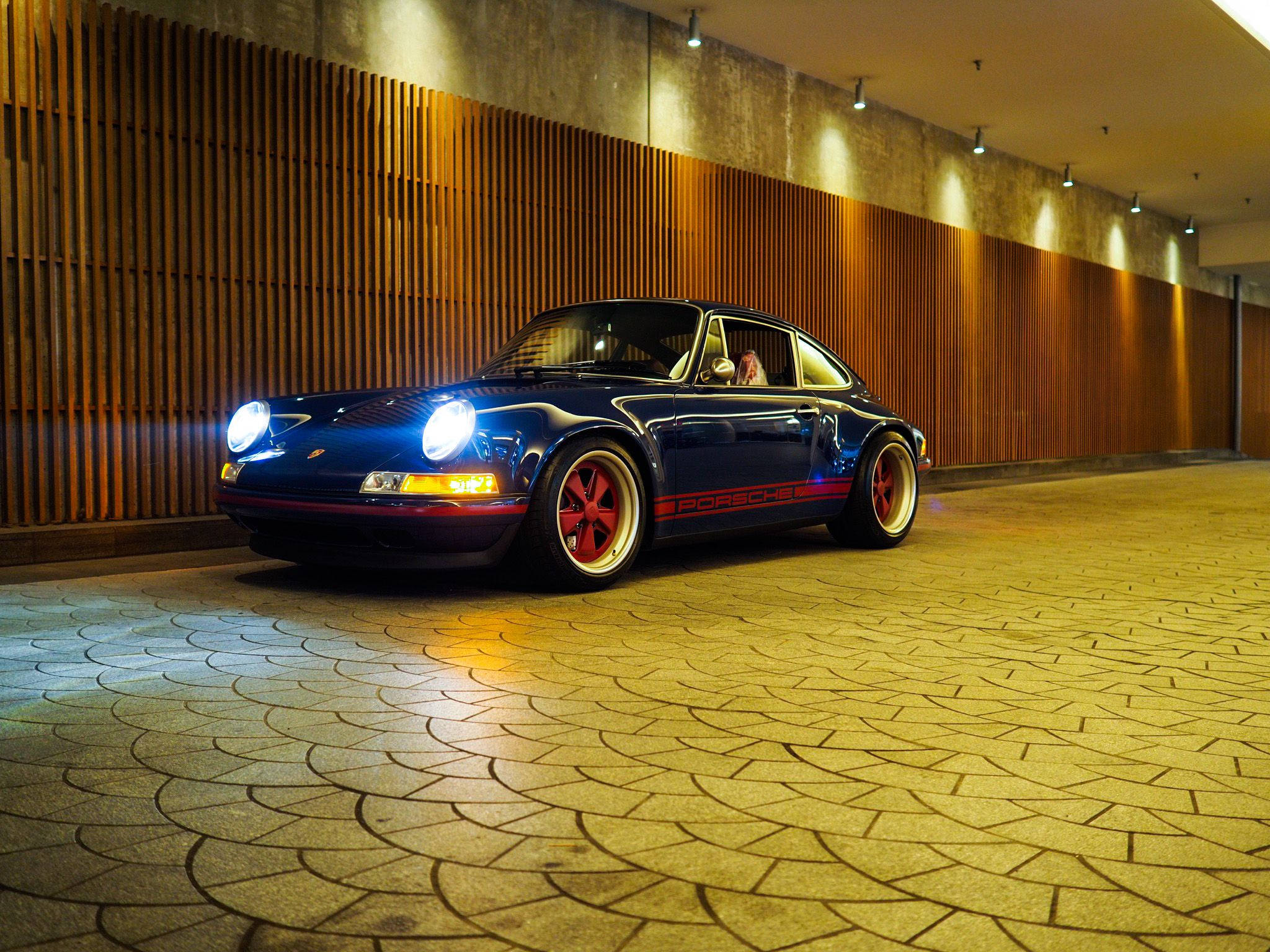 Singer 桌布 壁紙 — 一輛時尚的藍色保時捷911，帶有紅色點綴和發光的車頭燈，停在溫暖木條牆下的圖案石地板上。