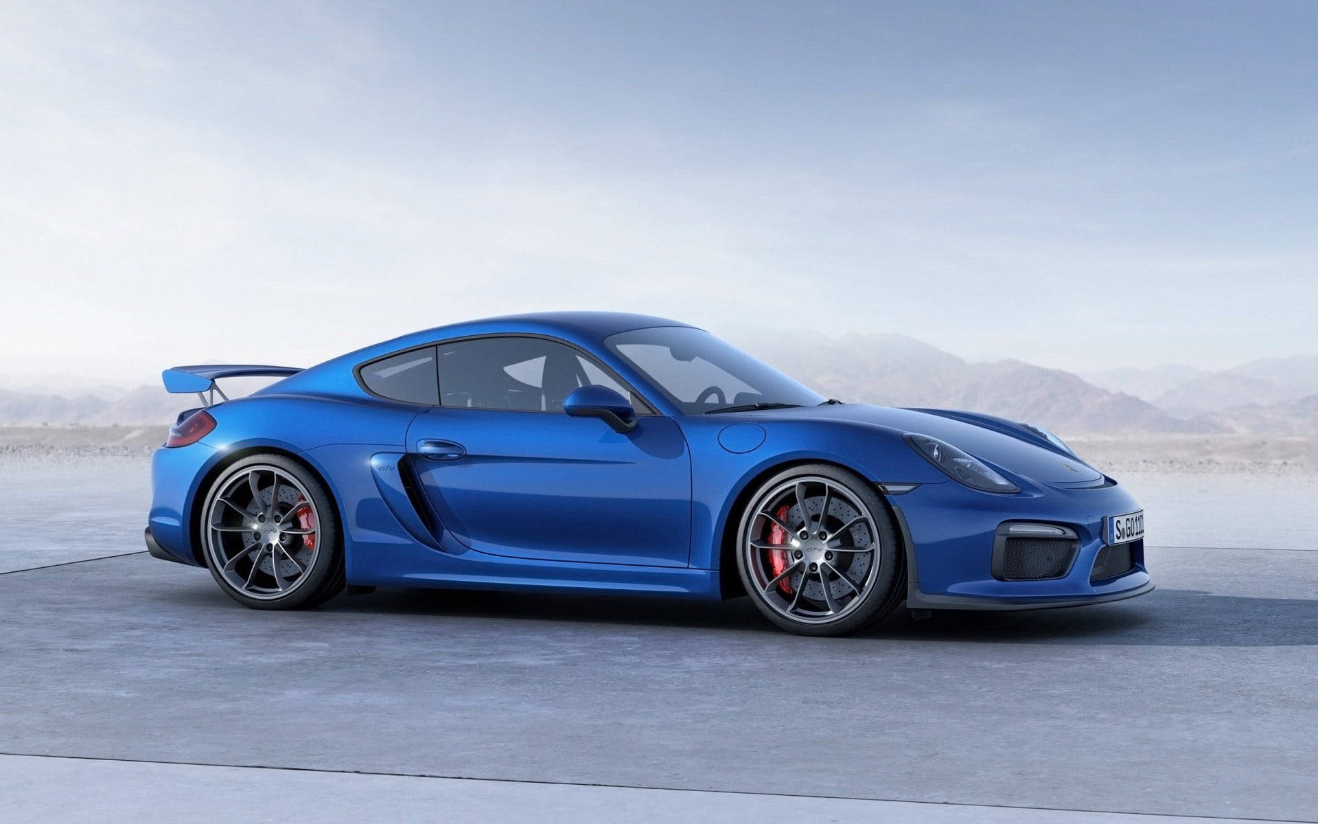 藍色 桌布 壁紙 — 一輛時尚的藍色保時捷Cayman GT4跑車，配備紅色制動卡鉗，停泊在混凝土平台上，背景是朦朧的山巒。