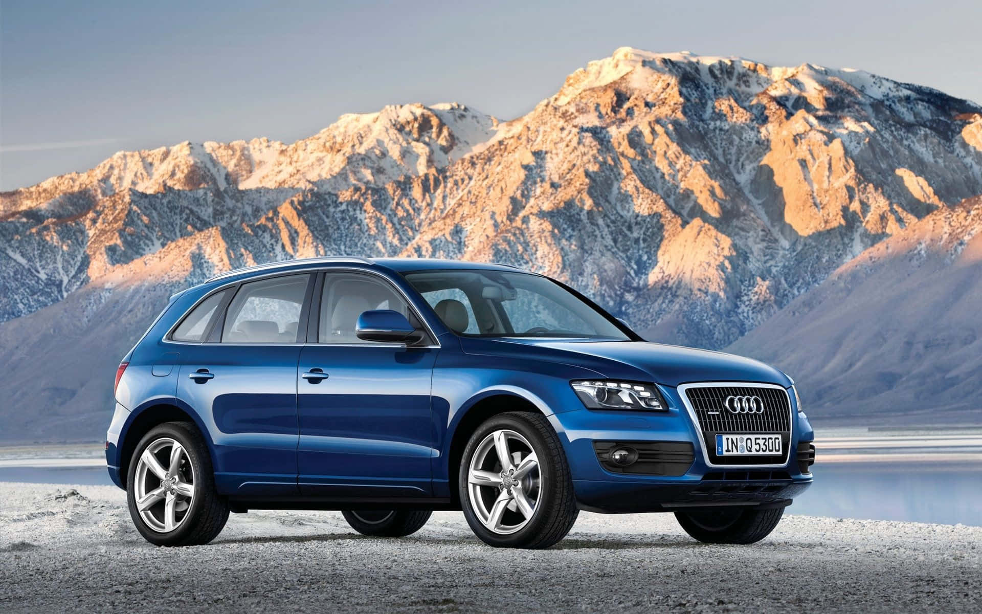 Audi Sq5 桌布 壁紙 — 一輛時尚的藍色奧迪Q5 SUV停在碎石路上，背後是積雪的山峰在金色夕陽下閃耀。