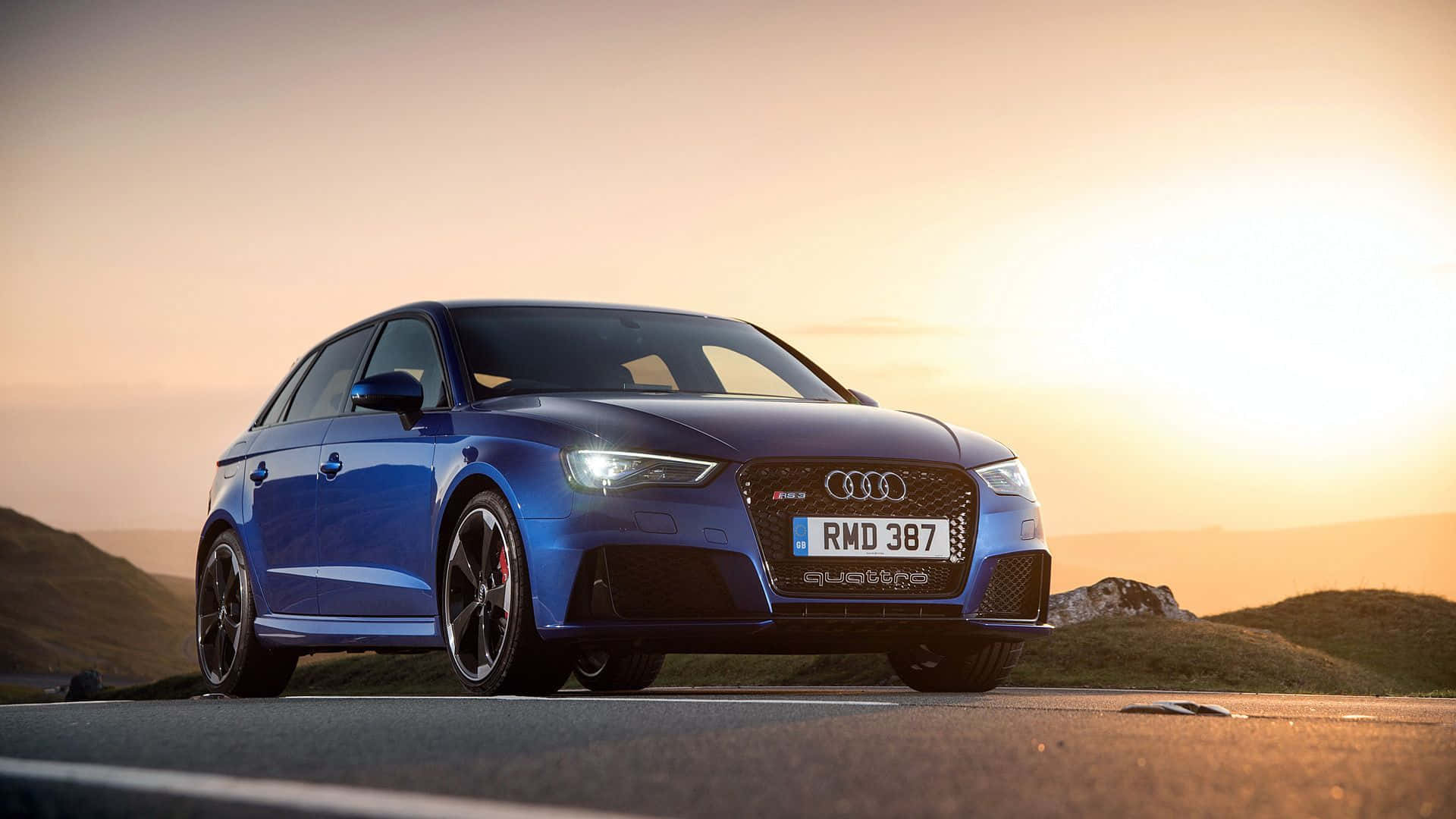 Audi Rs3 桌布 壁紙 — 一輛時尚的藍色奧迪RS3 Sportback在金色日落時分蜿蜒的道路上閃耀，背景是起伏的丘陵。