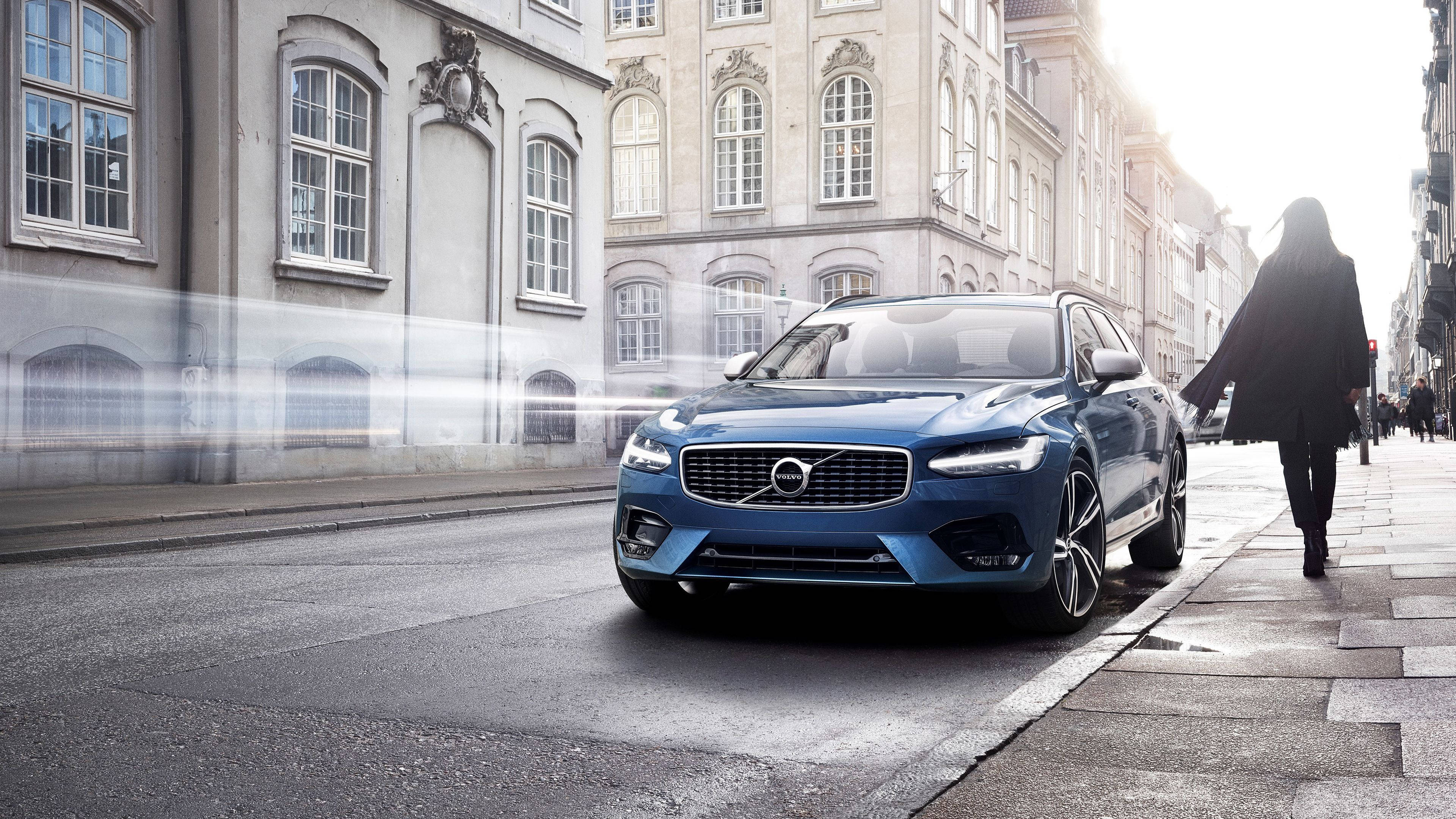 Volvo 桌布 壁紙 — 一輛時尚的藍色富豪SUV停在歐洲街道上，一名女子在明亮的陽光下以剪影走過。