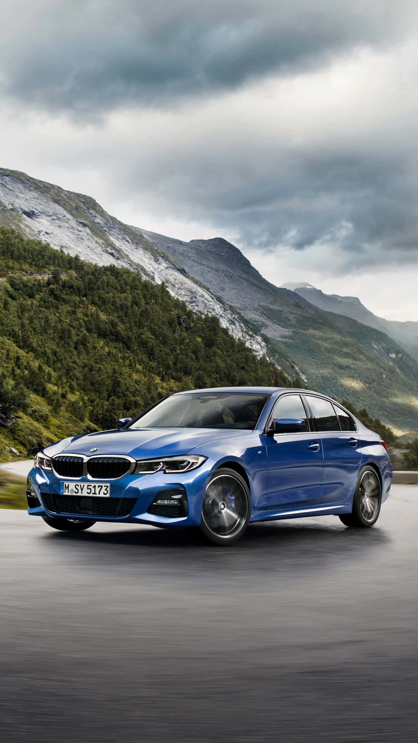 Bmw 328 桌布 壁紙 — 一輛時尚的藍色寶馬轎車在戲劇性的多雲天空下沿著山路行駛，背景是森林覆蓋的山峰。