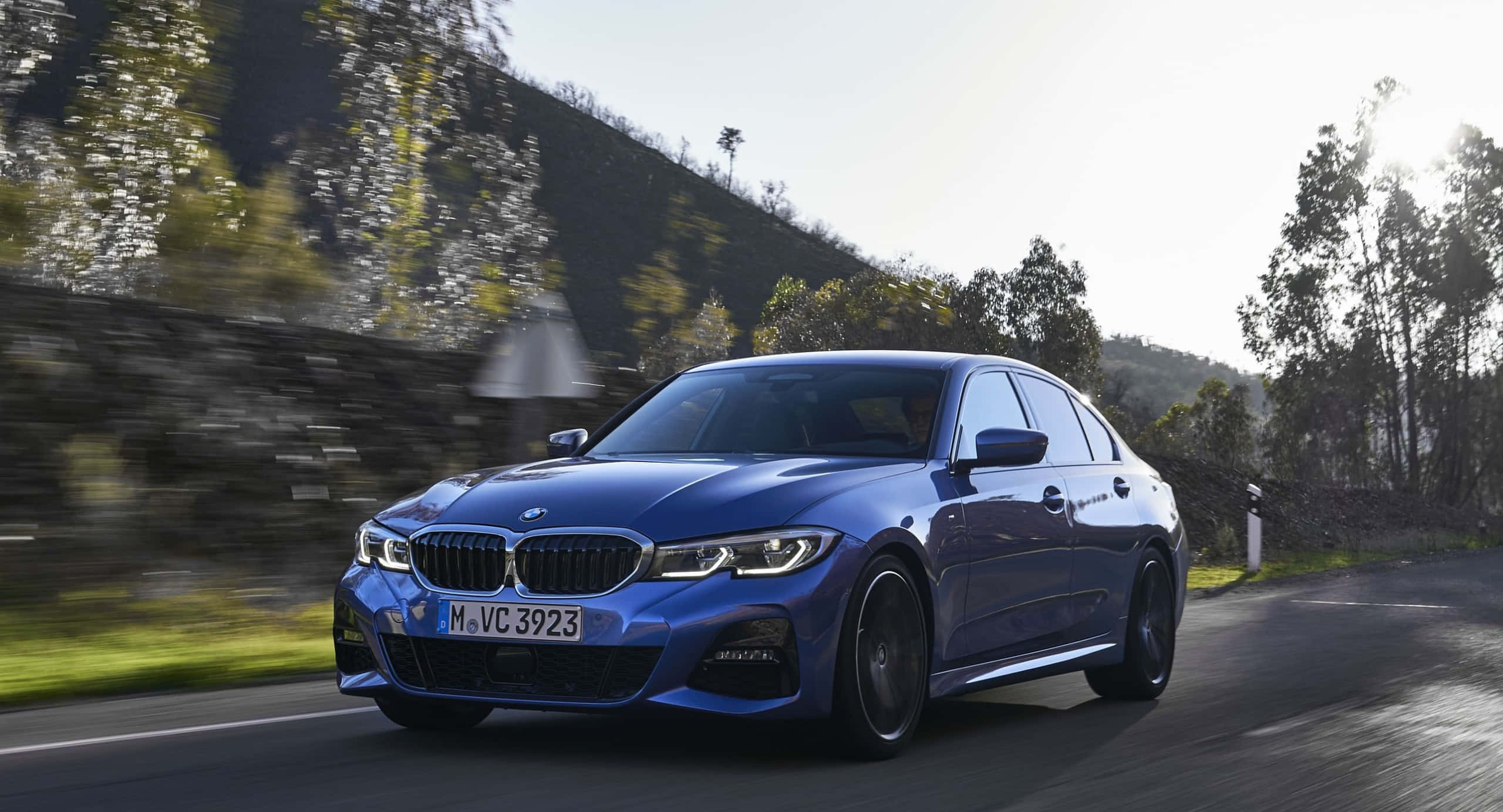 BMW 動圖 桌布 壁紙 — 一輛時尚的藍色寶馬轎車在明亮的日光下，沿著蜿蜒的山路疾馳，綠樹和石牆模糊成一片。