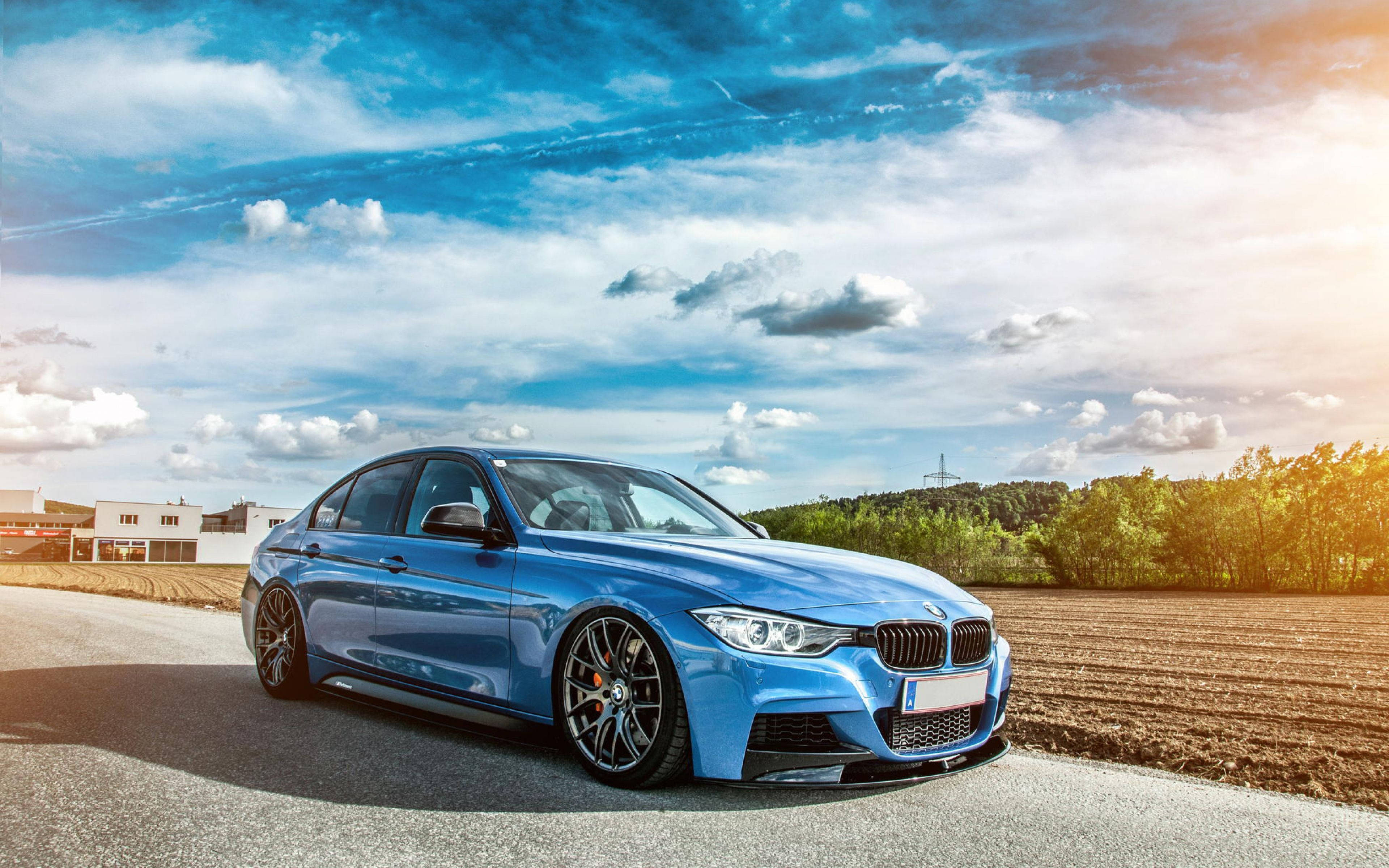 4K BMW 壁紙 桌布 壁紙 — 一輛時尚的藍色寶馬轎車在鄉村道路上，在充滿活力的天空和散落的雲朵下閃閃發光。
