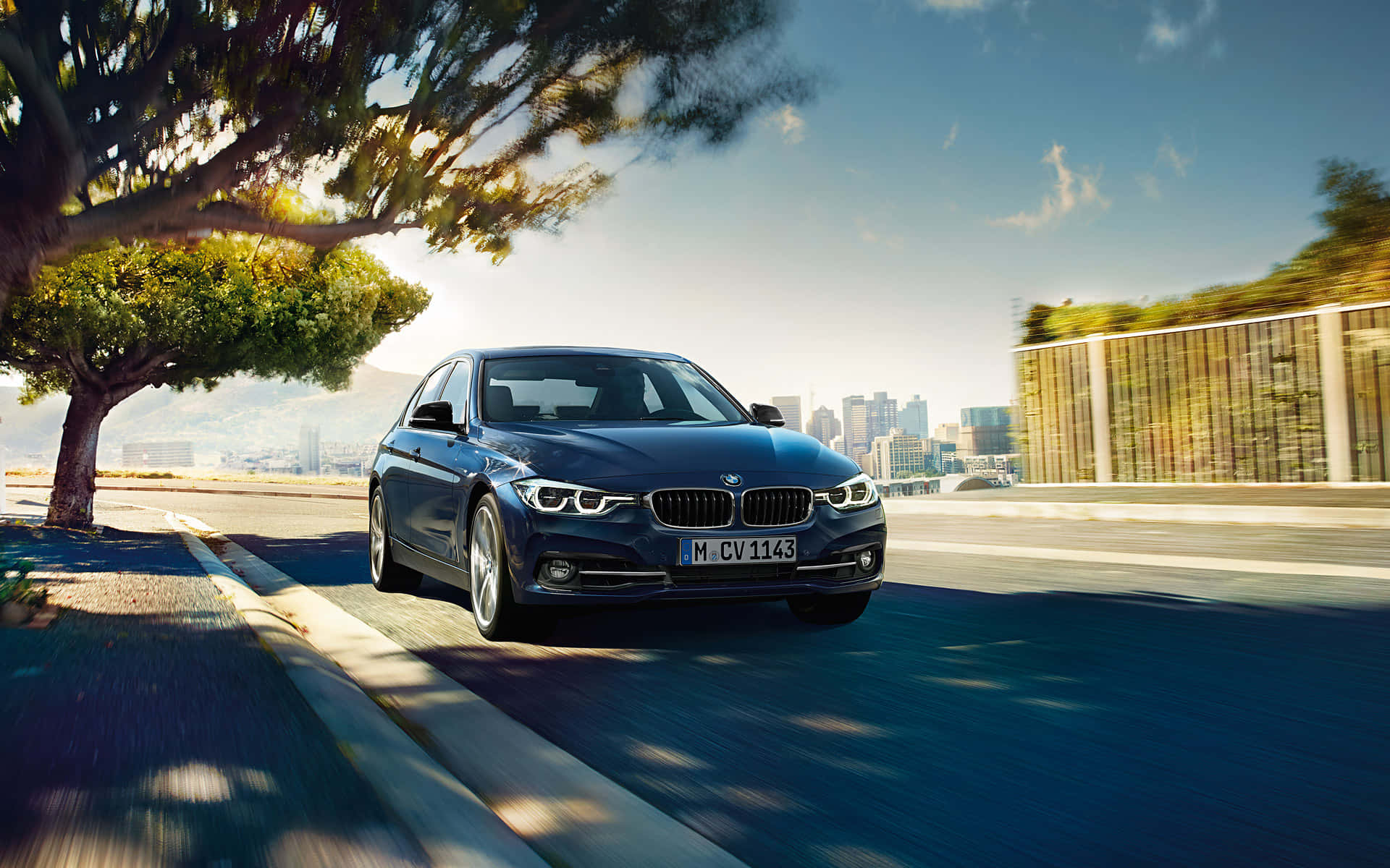Bmw 328 桌布 壁紙 — 一輛時尚的藍色寶馬轎車沿著陽光明媚的海岸道路行駛，背景是城市天際線和一棵枝葉茂密的樹木。