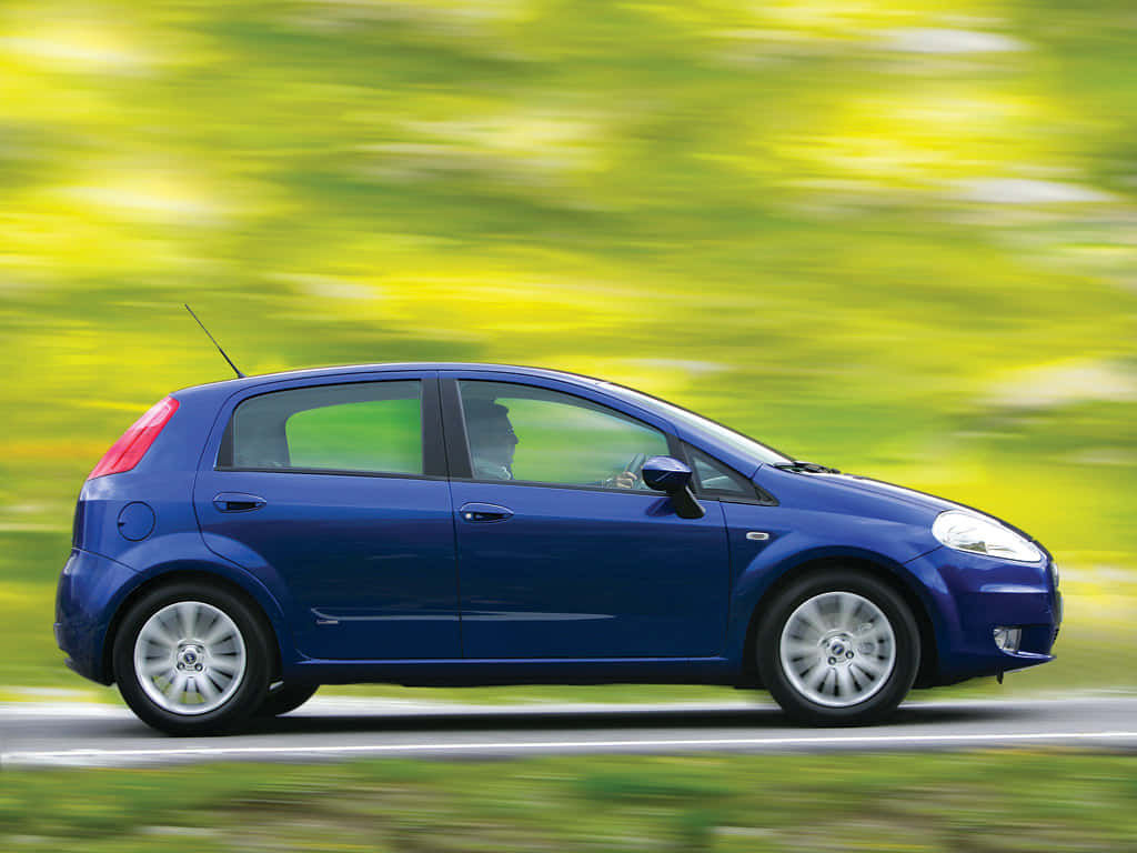 Fiat Punto 桌布 壁紙 — 一輛時尚的藍色掀背車在道路上行駛，充滿活力的綠色樹葉在動態模糊中掠過。