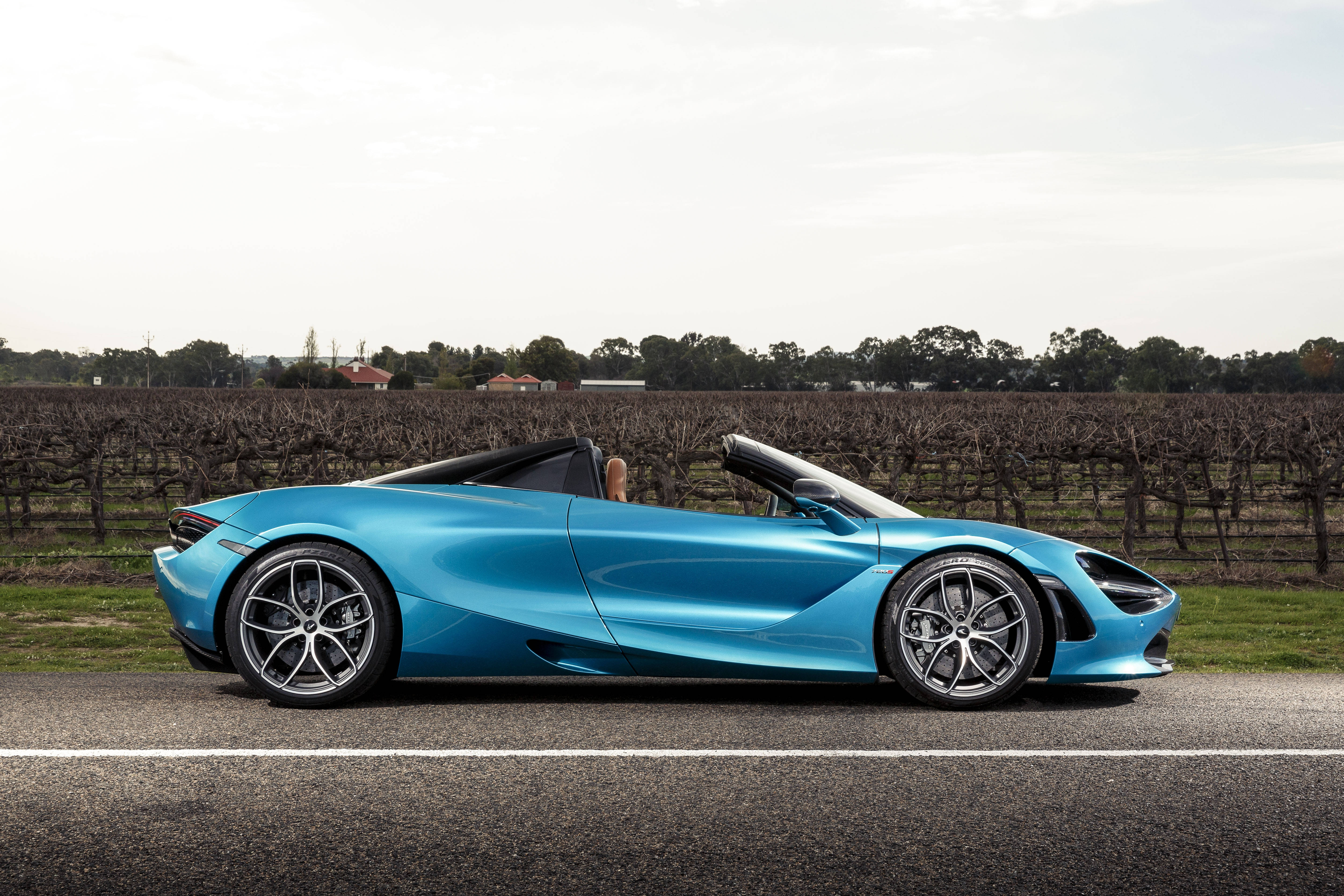 Mclaren 720s 桌布 壁紙 — 一輛時尚的藍色敞篷跑車在陰天下行駛在葡萄園旁邊的鄉村道路上。