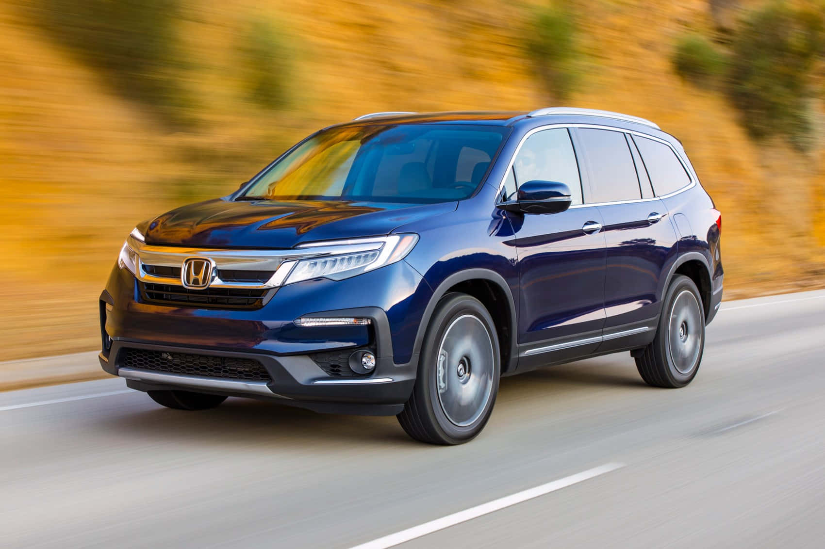 Honda Pilot 桌布 壁紙 — 一輛時尚的藍色本田 Pilot SUV 沿著蜿蜒的道路疾馳，背景是模糊的金色樹葉。