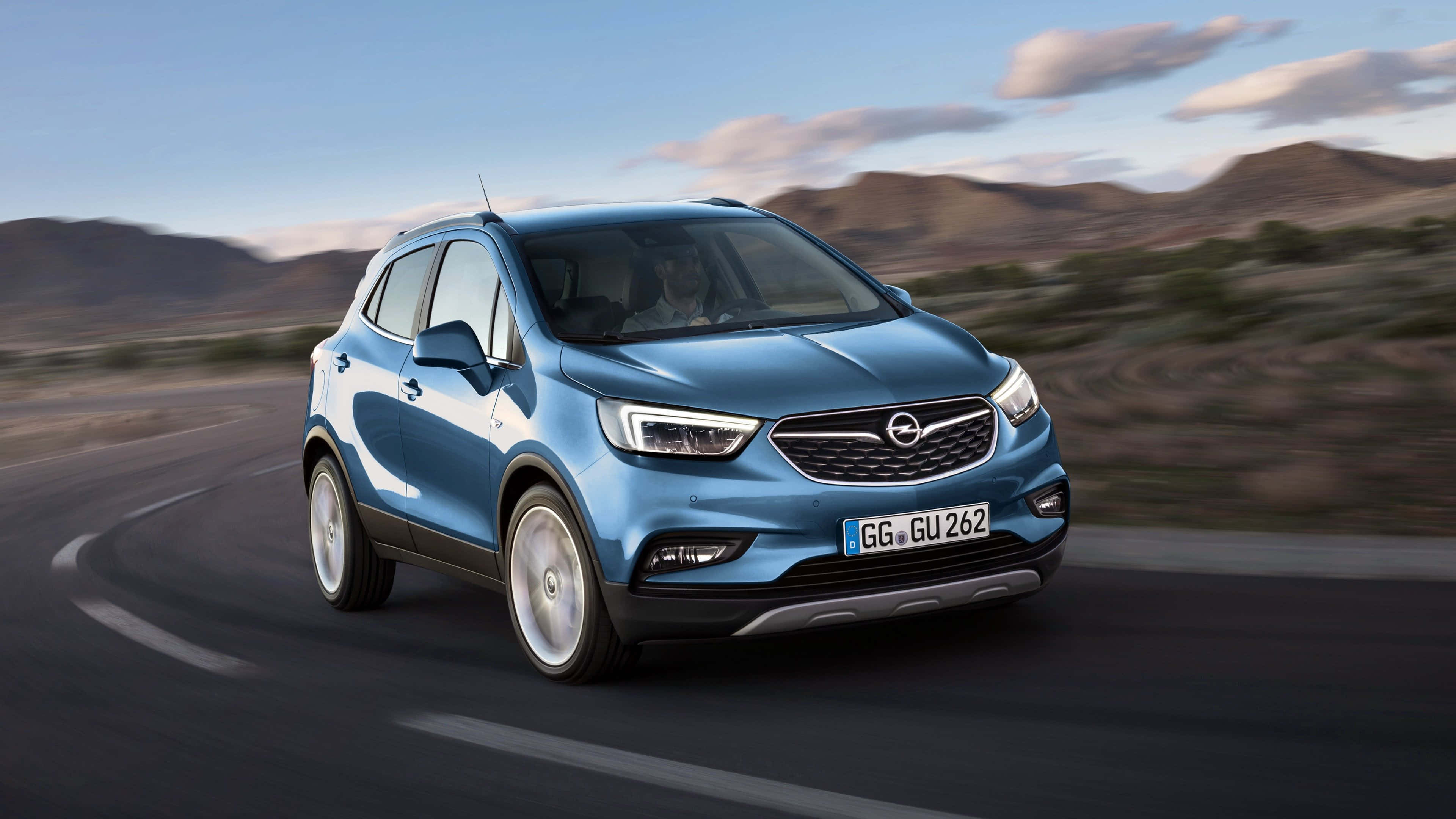 Opel Mokka 桌布 壁紙 — 一輛時尚的藍色歐寶 Mokka SUV 在柔和的暮色天空下沿著蜿蜒的沙漠公路疾馳。
