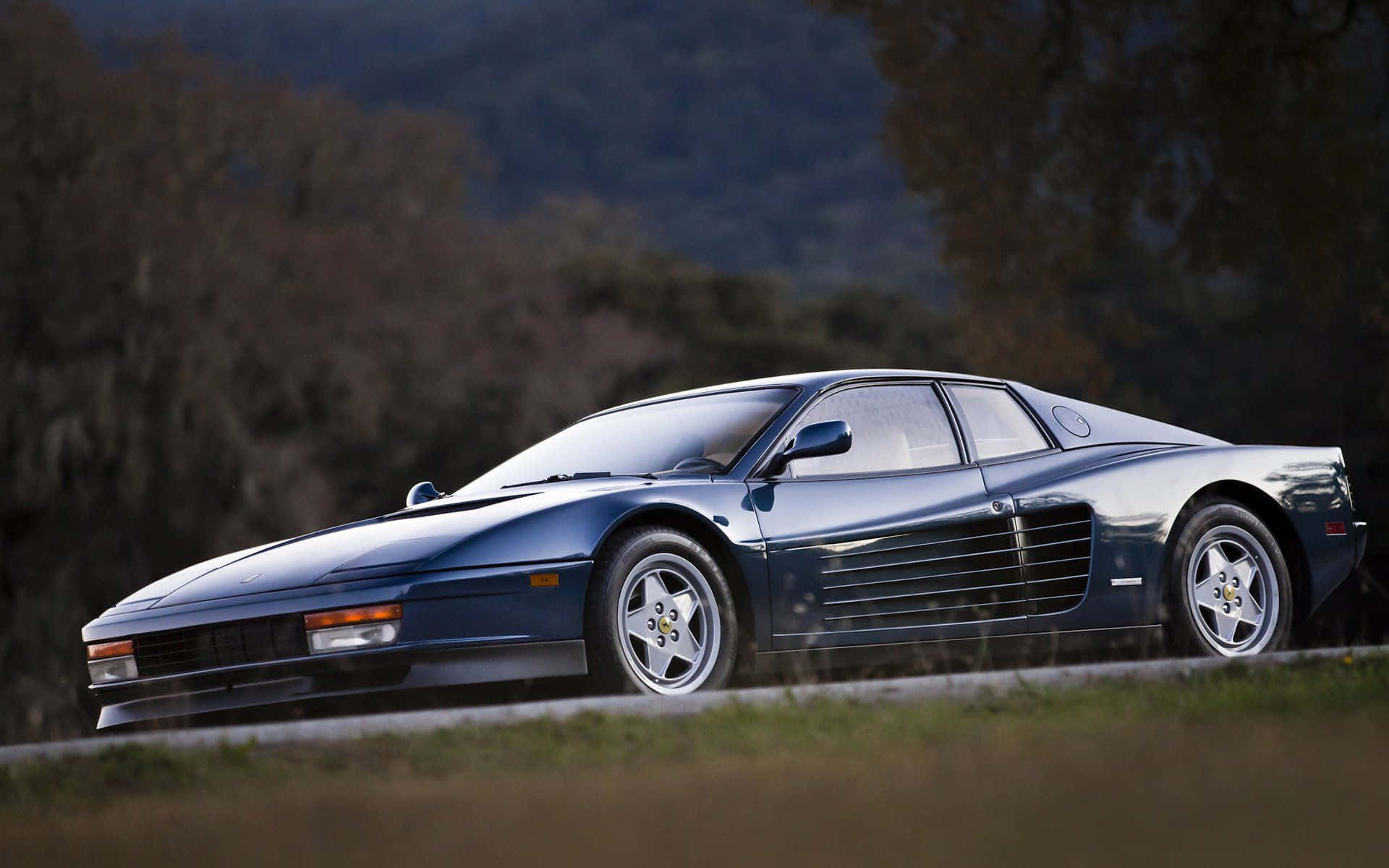 Ferrari Testarossa 桌布 壁紙 — 一輛時尚的藍色法拉利 Testarossa 在黃昏時分沿著蜿蜒的道路穿過森林覆蓋的山坡疾馳。