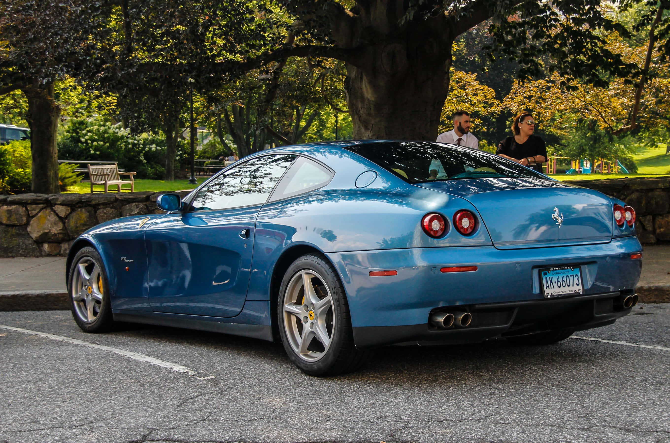 Ferrari 612 Scaglietti 桌布 壁紙 — 一輛時尚的藍色法拉利停泊在樹木林立的公園裡，附近有幾個人坐在陽光普照的日子裡。
