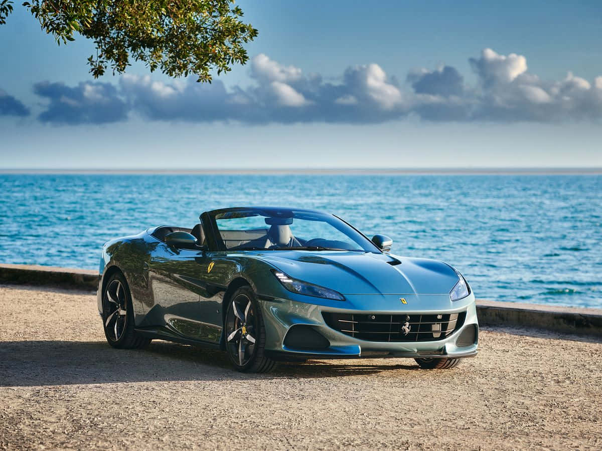 Ferrari Portofino 桌布 壁紙 — 一輛時尚的藍色法拉利敞篷車停放在陽光明媚的海灘上，背景是碧綠的海洋和散落的雲朵。