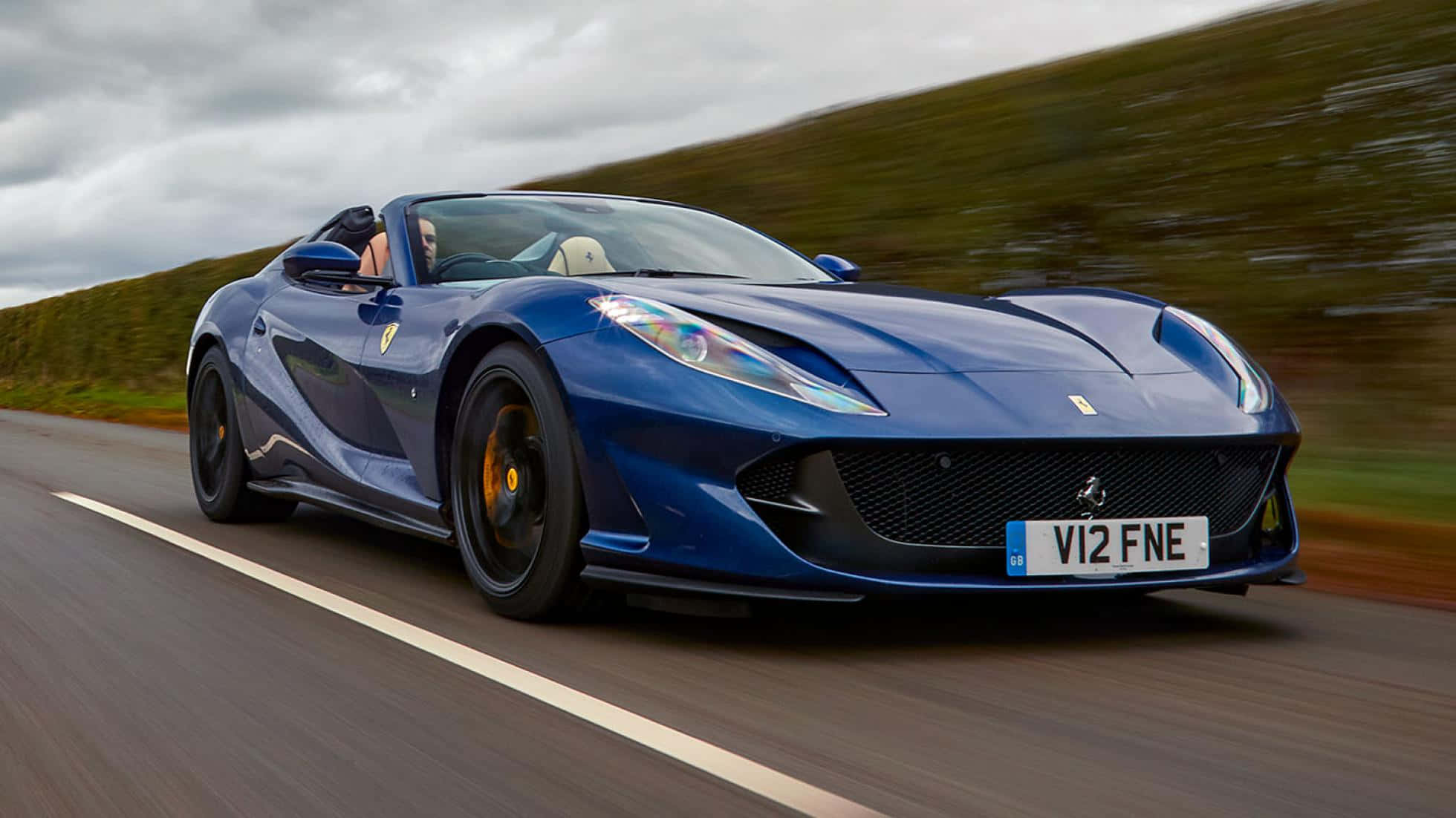 Ferrari 812 Superfast 桌布 壁紙 — 一輛時尚的藍色法拉利跑車在蜿蜒的道路上疾馳，背景是模糊的綠色山丘。