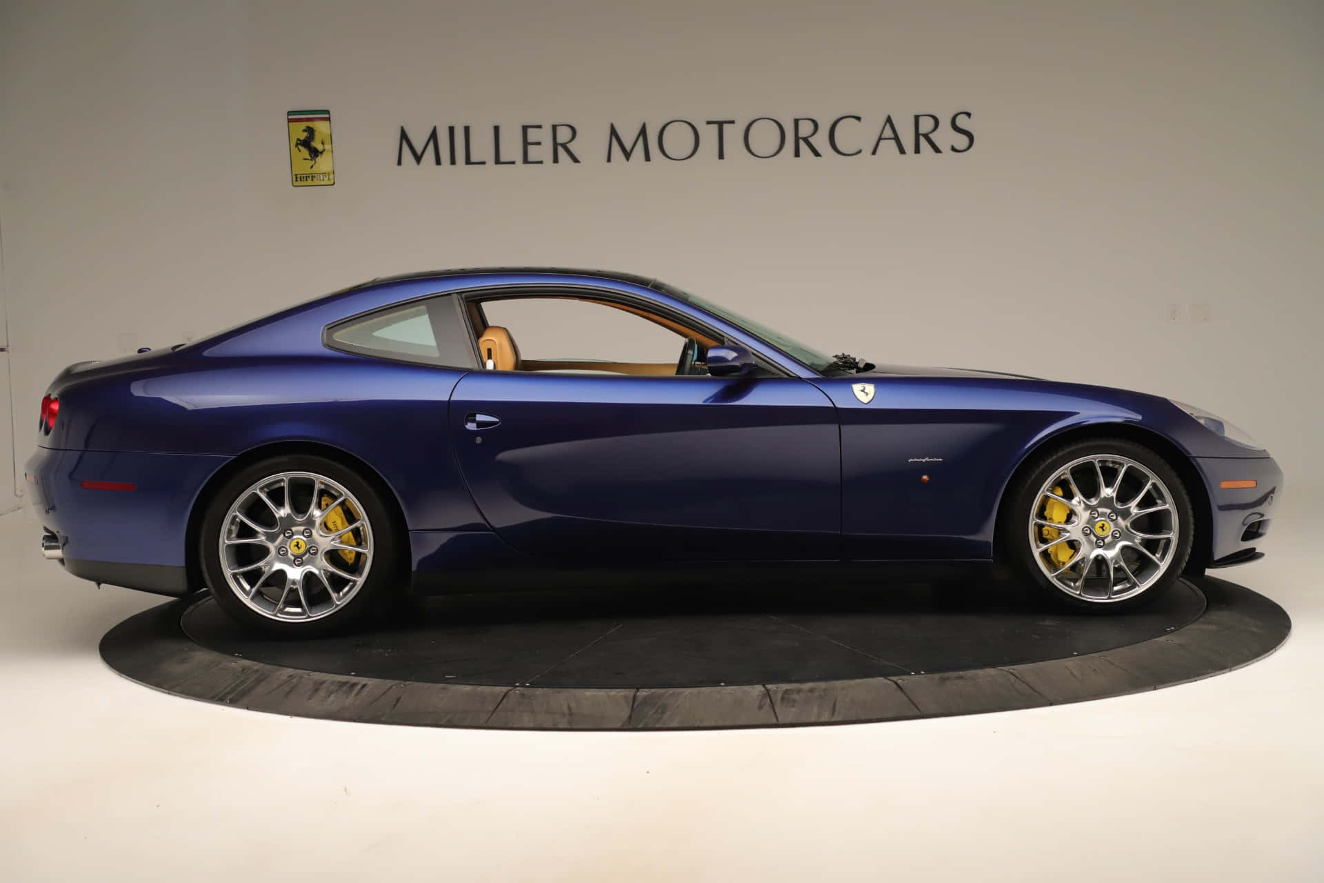 Ferrari 612 Scaglietti 桌布 壁紙 — 一輛時尚的藍色法拉利跑車配備黃色制動卡鉗，在純白色展廳的圓形平台上展示。