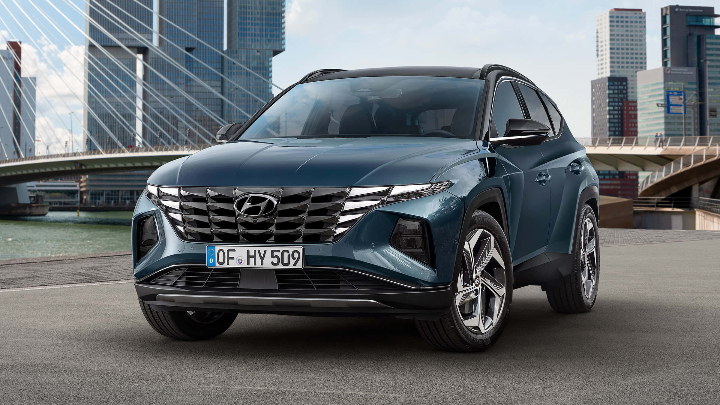 Hyundai 桌布 壁紙 — 一輛時尚的藍色現代 Tucson SUV 停泊在都市街道上，背景是現代摩天大樓和斜張橋。