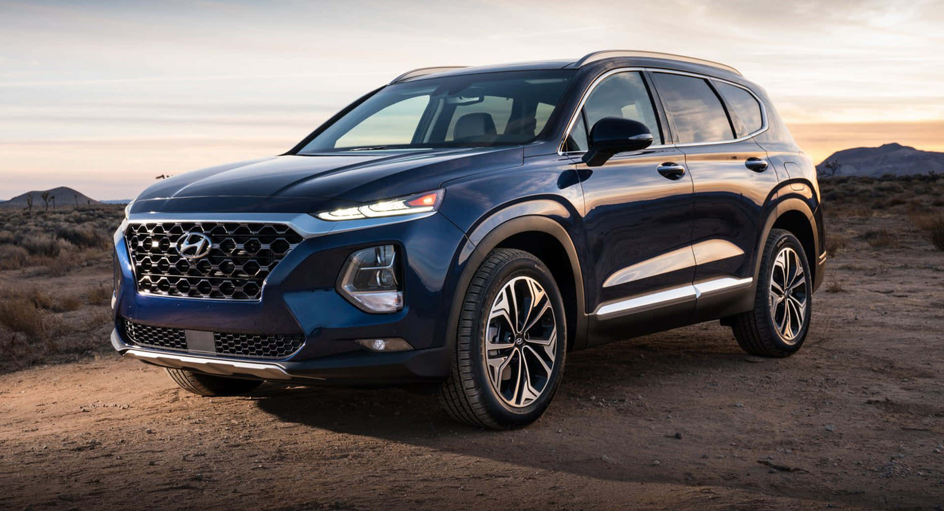 Hyundai 桌布 壁紙 — 一輛時尚的藍色現代Santa Fe SUV停泊在塵土飛揚的地形上，背景是溫暖的日落天空和遠山。