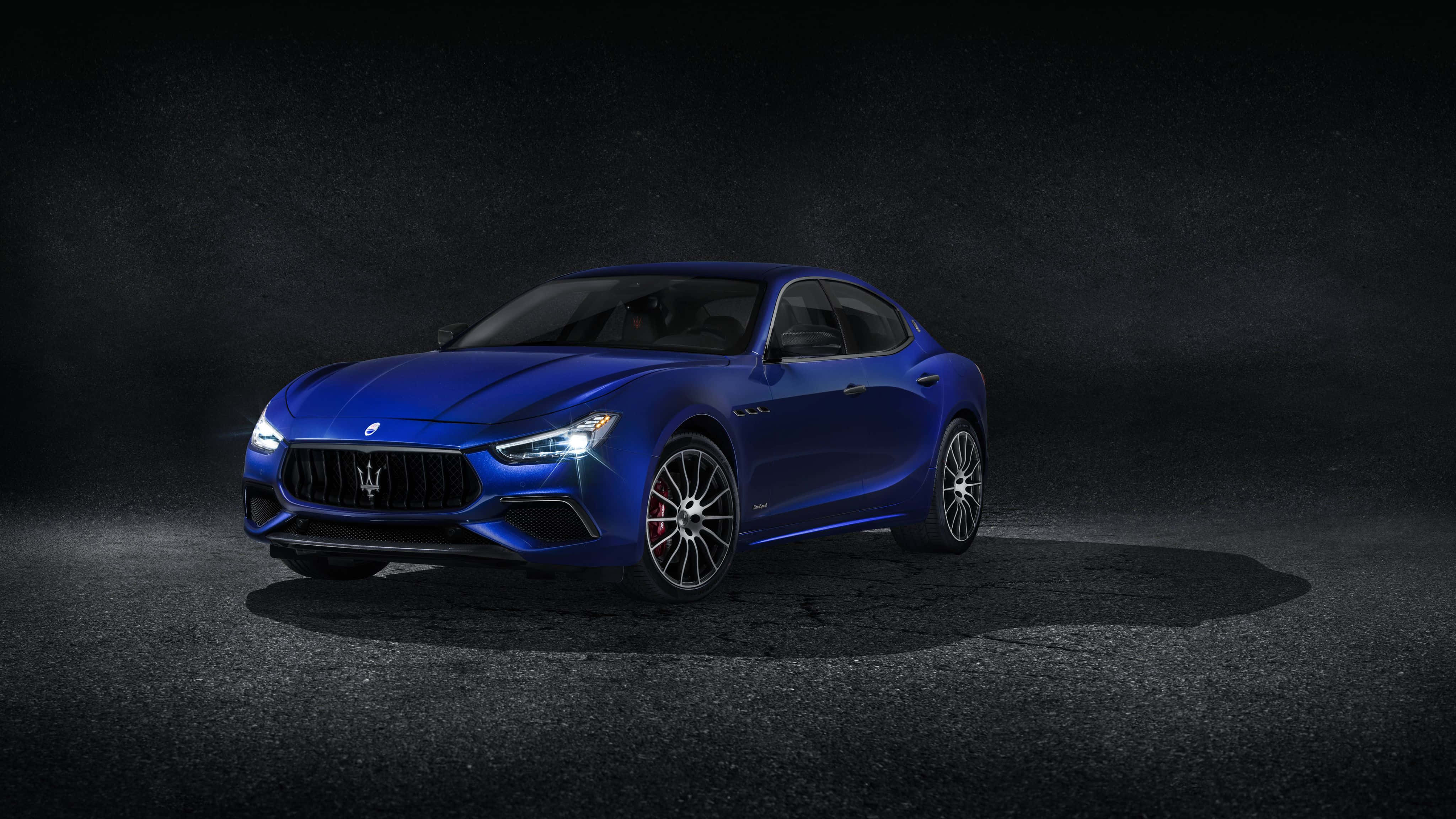 Maserati Ghibli 桌布 壁紙 — 一輛時尚的藍色瑪莎拉蒂跑車在戲劇性的聚光燈下，在深色紋理工作室背景前閃閃發光。