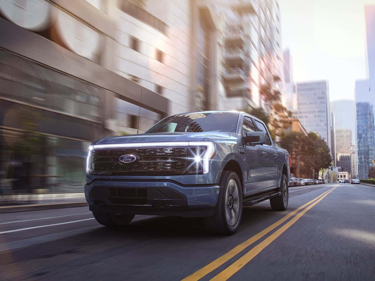 福特 桌布 壁紙 — 一輛時尚的藍色福特 F-150 Lightning 皮卡在陽光明媚的城市街道上奔馳，背景是動態模糊的摩天大樓。