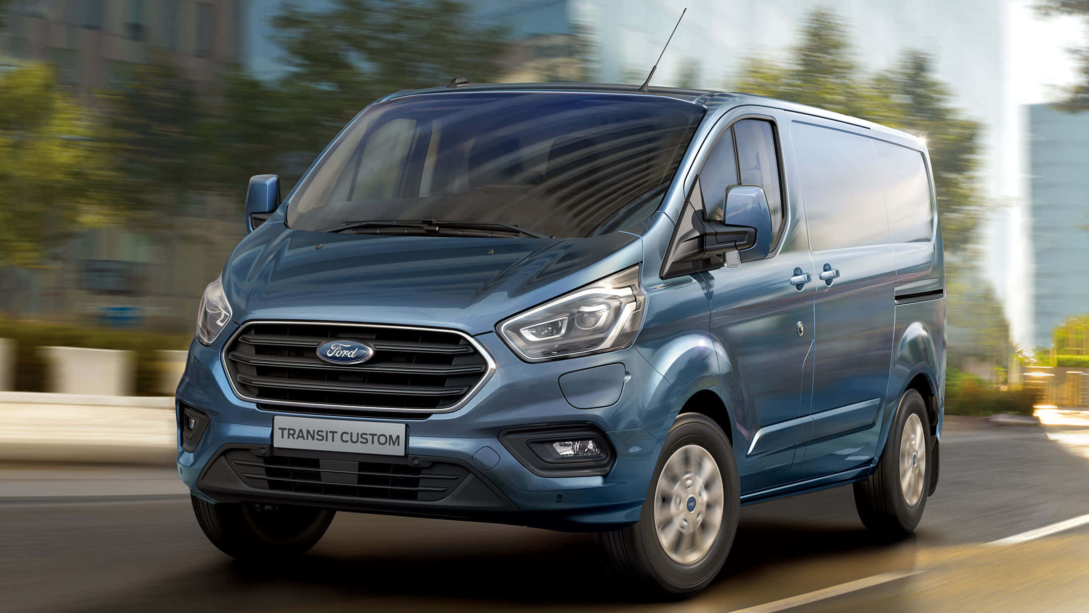 Ford Transit 桌布 壁紙 — 一輛時尚的藍色福特 Transit Custom 客貨車在模糊的城市背景下沿著城市街道飛馳。