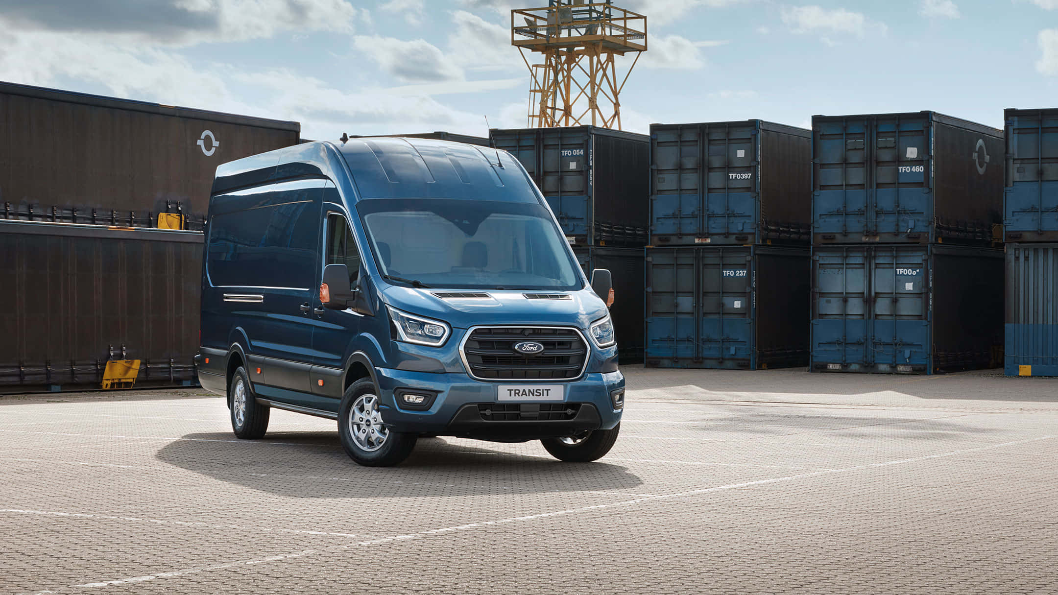 Ford Transit 桌布 壁紙 — 一輛時尚的藍色福特 Transit 貨車停放在鋪砌的庭院中，周圍堆疊著貨運集裝箱，天空部分多雲。