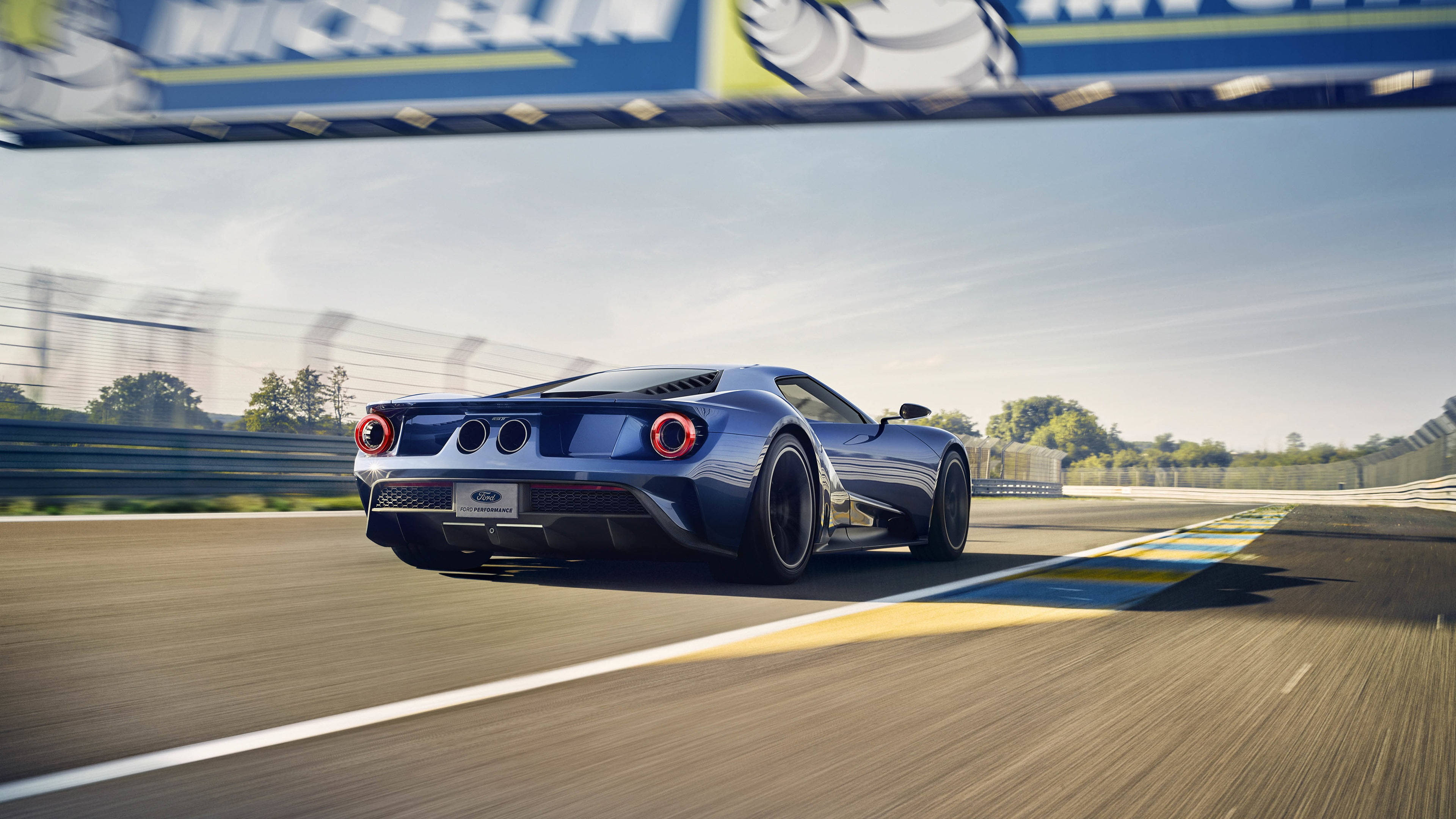 Racing 桌布 壁紙 — 一輛時尚的藍色福特GT在陽光明媚的賽道上奔馳，帶有動態模糊效果，上方有米其林橫幅。