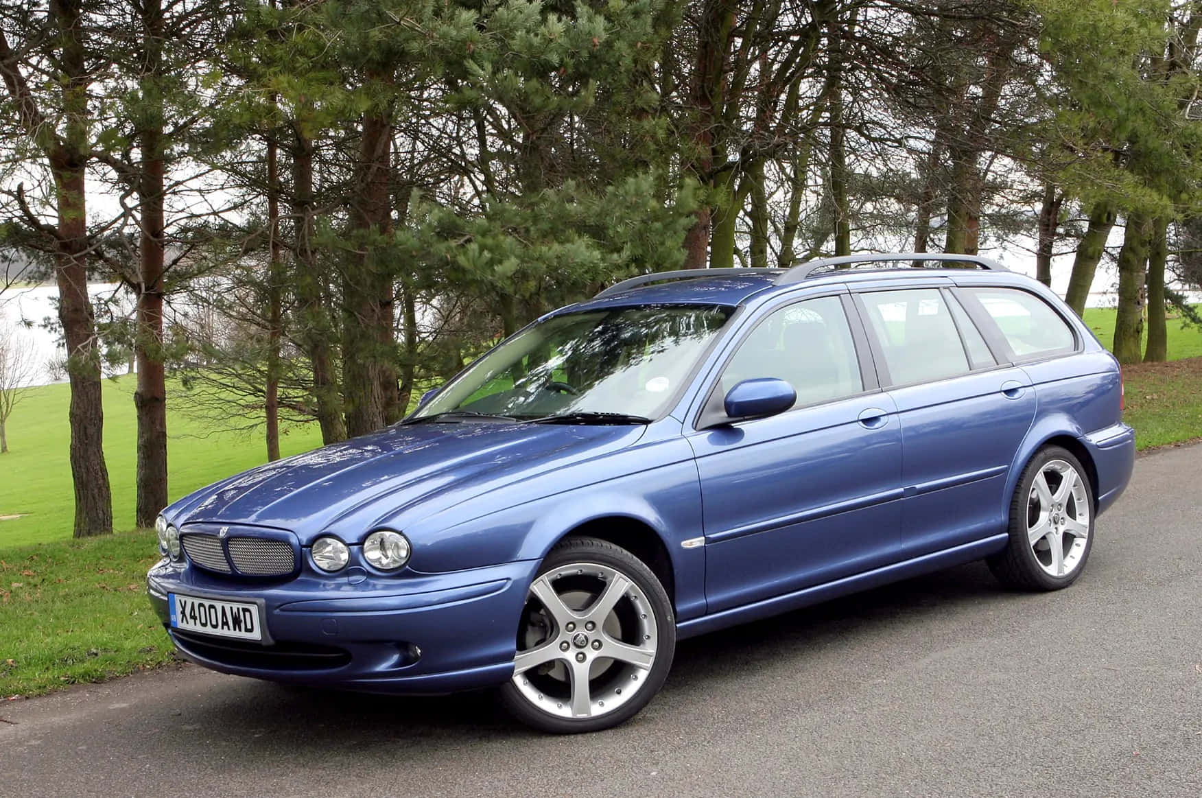 Jaguar X-type 桌布 壁紙 — 一輛時尚的藍色積架旅行車停泊在鋪砌的道路上，旁邊有綠草和高大的松樹。