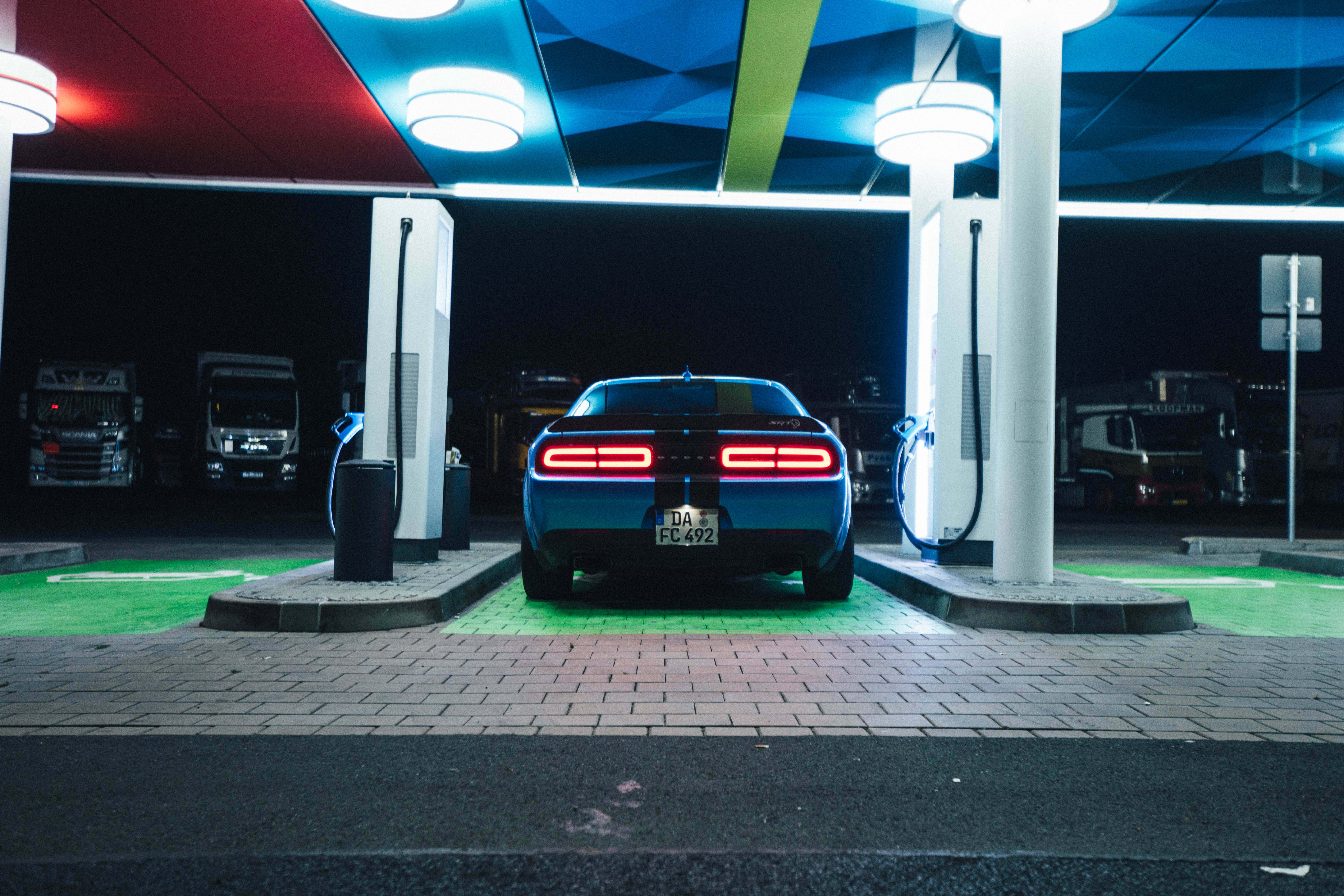 Challenger 桌布 壁紙 — 一輛時尚的藍色肌肉車在夜間充滿活力的霓虹燈下於現代電動站充電。