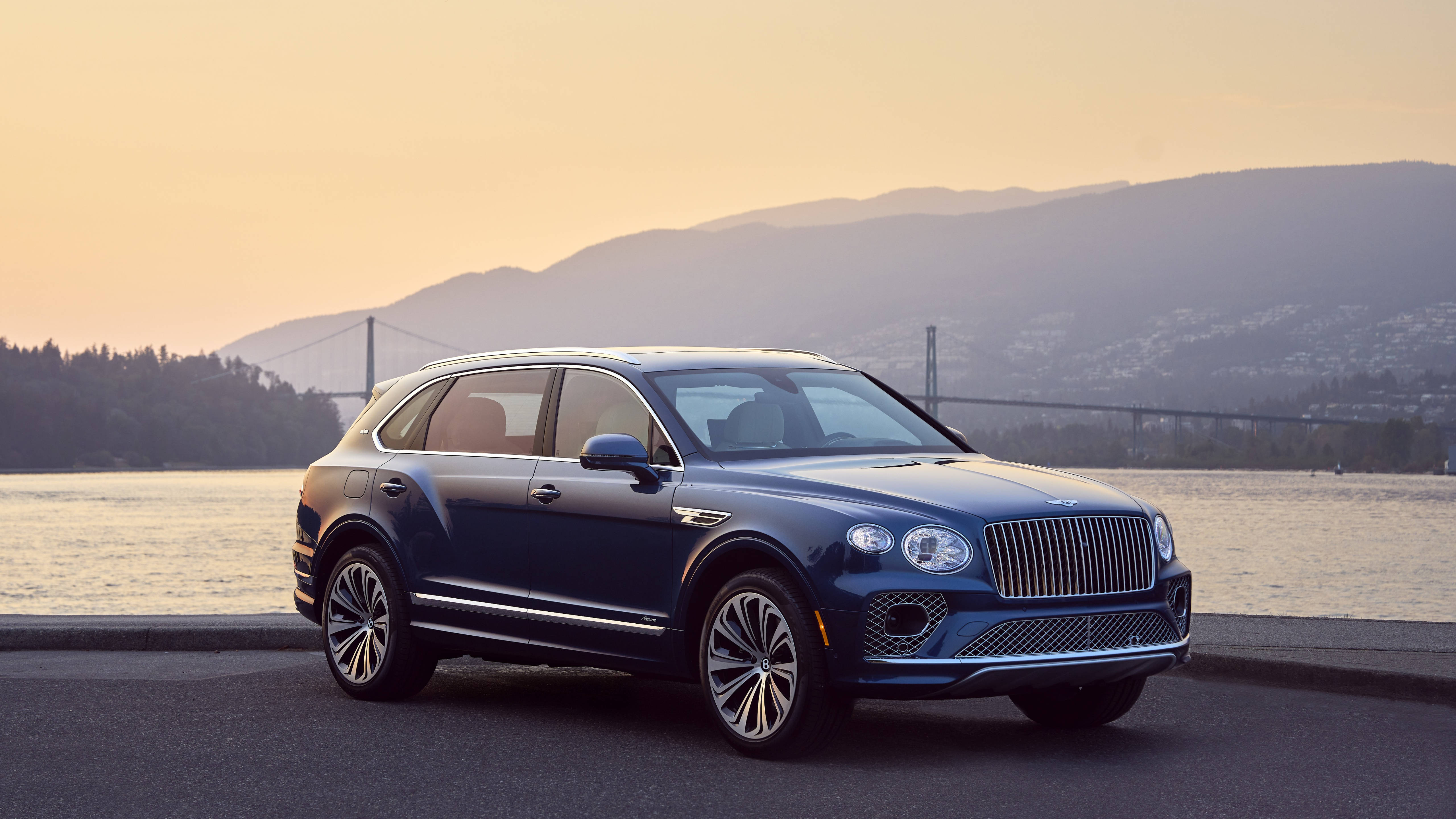 bentley HD 桌布 壁紙 — 一輛時尚的藍色賓利 Bentayga SUV 停泊在水邊，背景有山脈和橋樑，沐浴在金色的日落餘暉中。