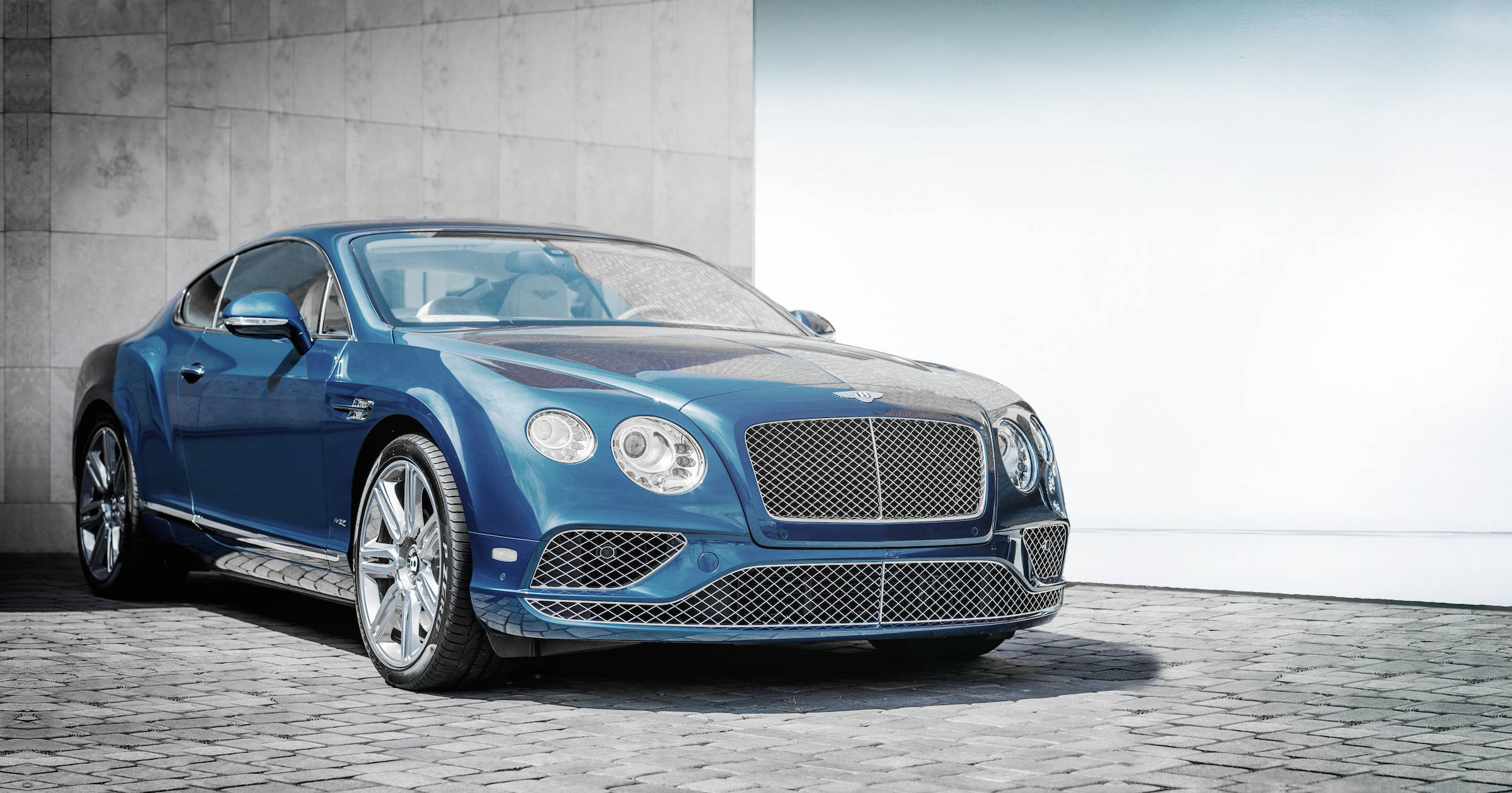 cars 桌布 壁紙 — 一輛時尚的藍色賓利 Continental GT 轎跑車在明亮光線下，以簡約的混凝土牆和鵝卵石地面為背景閃閃發光。