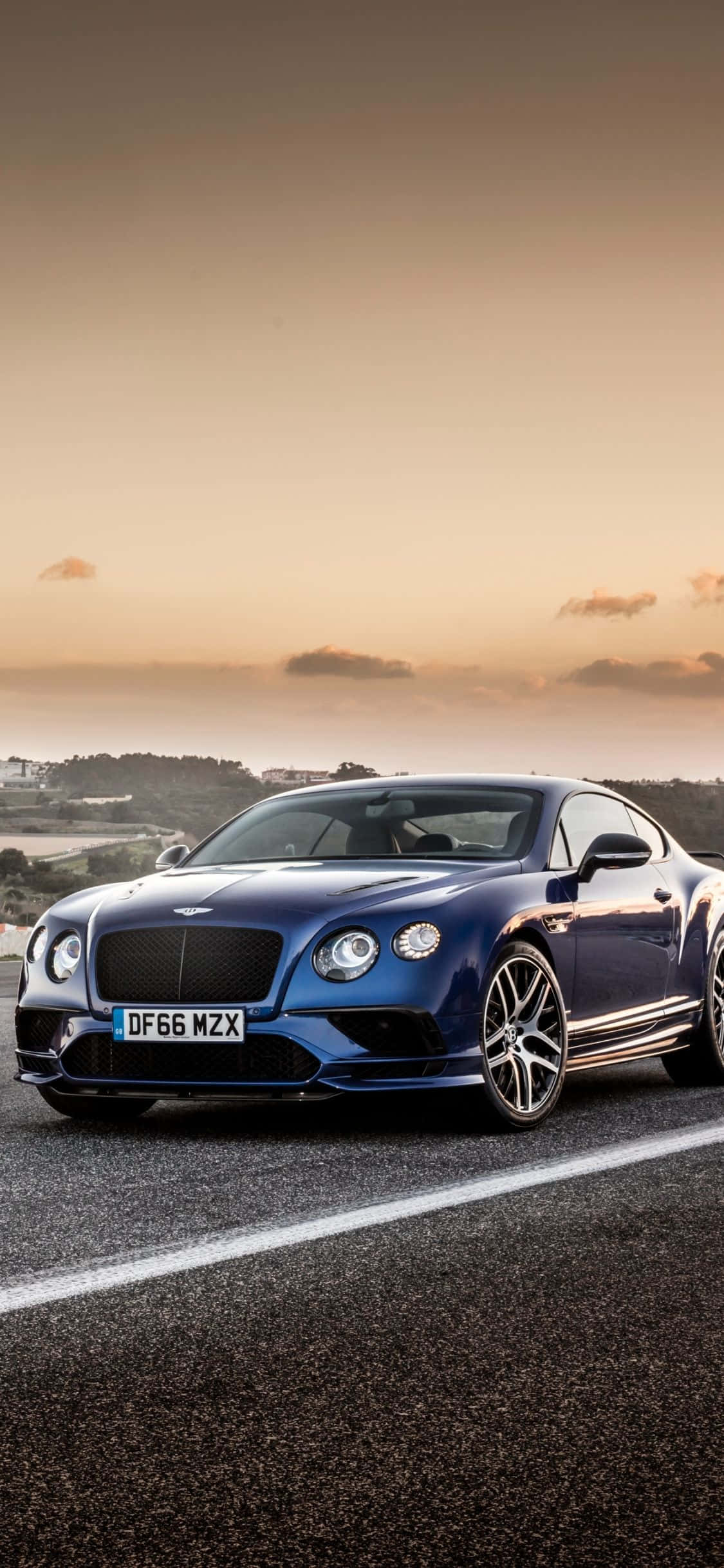 Blue Car 桌布 壁紙 — 一輛時尚的藍色賓利 Continental GT 在日落時分沿著海岸公路疾馳，伴隨著溫暖的金色光芒。