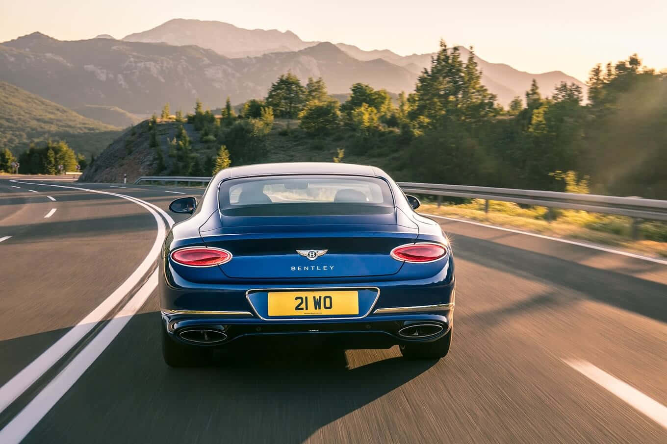 Convertible 桌布 壁紙 — 一輛時尚的藍色賓利 Continental GT 在黃金時段沿著蜿蜒的山區高速公路行駛，背景是森林覆蓋的山丘。