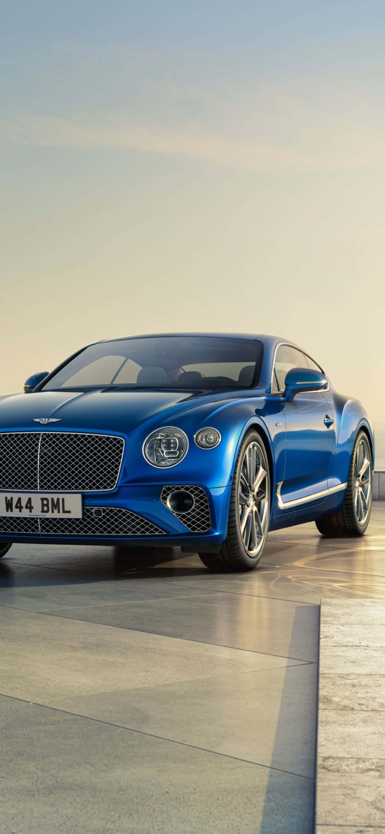 iPhone Xs Max Bentley 桌布 壁紙 — 一輛時尚的藍色賓利 Continental GT 雙門跑車在金色時刻的光線下，在拋光混凝土表面上閃閃發光。
