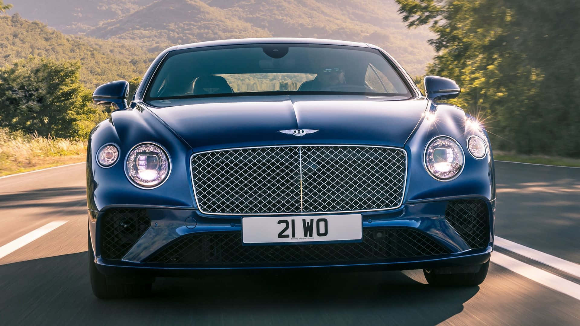Best Bentley 桌布 壁紙 — 一輛時尚的藍色賓利Bentayga在陽光明媚的山路上疾馳，背景是茂密的綠植和遠山。