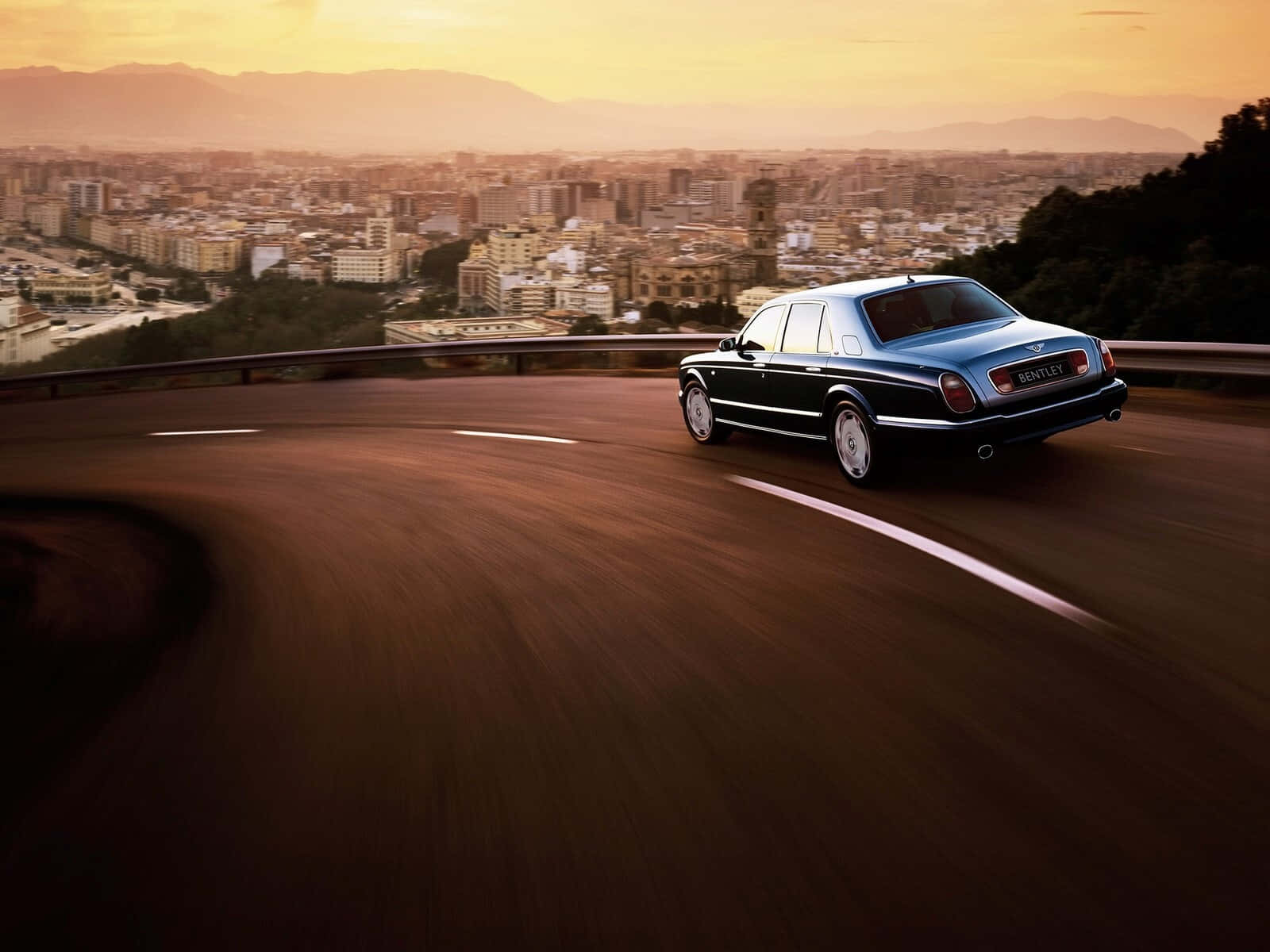 Bentley Arnage 桌布 壁紙 — 一輛時尚的藍色賓利在蜿蜒的山路上疾馳，俯瞰著金色日落下一座廣闊的城市。